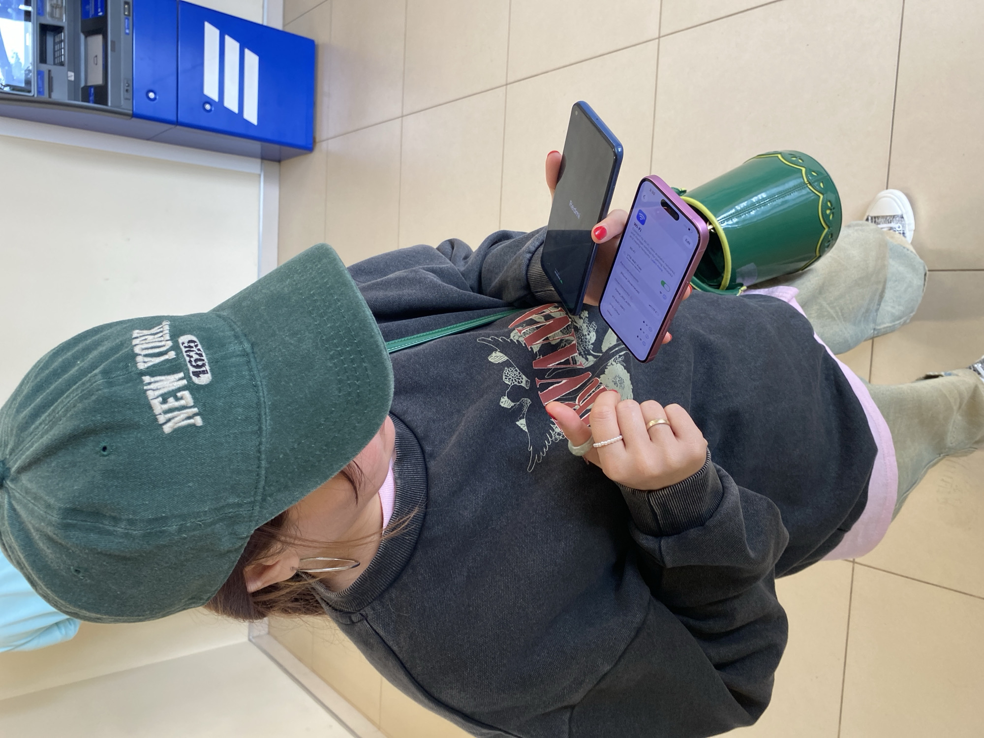 A person wearing a dark green baseball cap with "NEW YORK 1625" embroidered on it stands in front of a blue ATM. They are also wearing a dark grey sweatshirt over a pink shirt, light-colored pants, and white sneakers. The person is holding two smartphones in their hands, one with a pink case and the other black. The background shows a light-colored wall and tiled floor. The image appears to have been taken indoors, possibly in a public area like a bank lobby. The city of Votkinsk, Russia is not visibly identifiable in the image.