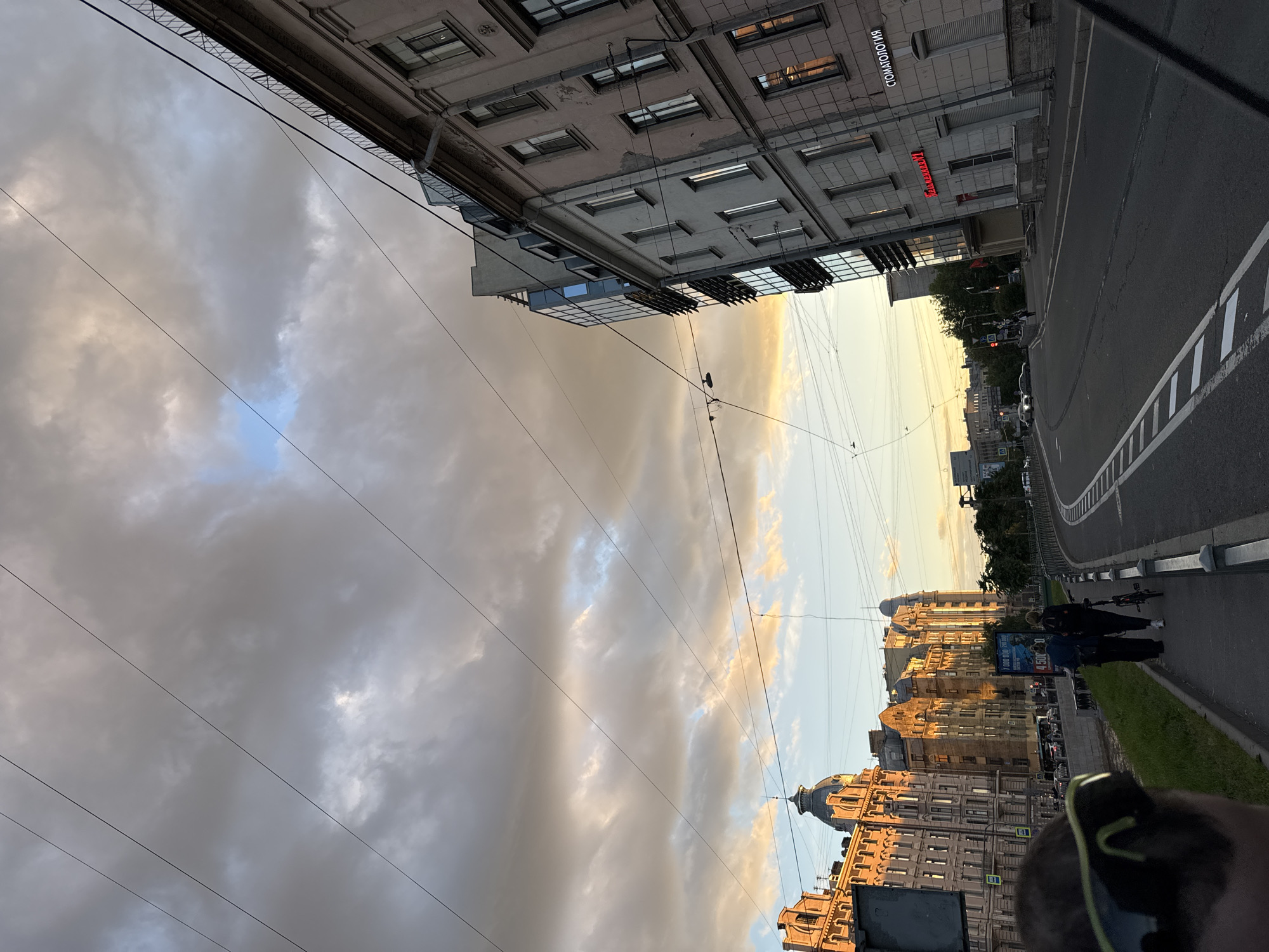The image captures a street scene in Saint Petersburg, Russia, likely during the late afternoon or early evening, given the warm, golden light illuminating the buildings. The sky is filled with dramatic, textured clouds in shades of gray, white, and hints of yellow and blue. Power lines crisscross the sky, a common feature of urban landscapes.
On the left side of the street, ornate, historical buildings with distinct architectural details and domed roofs are bathed in sunlight. A canal runs alongside the sidewalk. On the right, a series of buildings, some appearing more modern with glass facades, line the street. One building has a neon sign in Russian reading "СТОМАТОЛОГИЯ" (Dentistry). Another sign in red letters reads "РЕСТОРАН" (Restaurant).
Several people are visible walking along the sidewalk. Two individuals, one with a backpack and another pushing a bicycle, are walking away from the camera. In the foreground, a person is partially visible from behind, wearing sunglasses, suggesting they are looking out at the scene. Cars are visible on the street, and traffic signals indicate an active urban environment. The road is paved and marked with white lines. The overall impression is of a vibrant city street at a beautiful time of day.