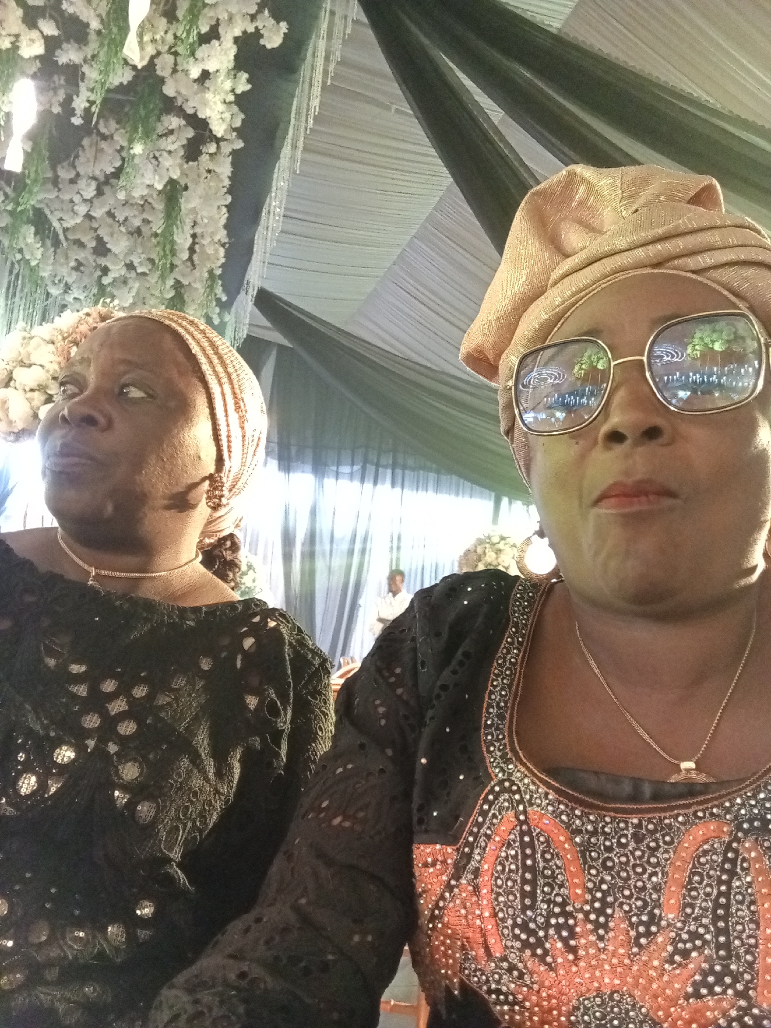 In Barden Yana, Nigeria, two women are posing for a photograph indoors. The woman on the left, with dark skin and brown eyes, wears a black lace dress and a light brown headscarf. She is looking to her left. The woman on the right, also with dark skin and brown eyes, wears a gold headscarf and large, gold-rimmed glasses. Her black dress is adorned with colorful embellishments around the neckline. She is looking directly at the camera. Both women are wearing necklaces. The background shows decorative white flowers hanging from above and draped fabric, suggesting a formal event or celebration.