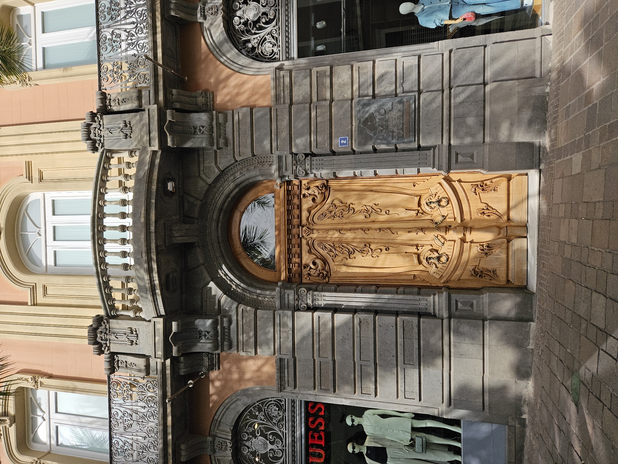 The image is an eye-level shot of the architectural facade of a building in Santa Cruz De Tenerife, Spain. The primary focus is on a large, ornately carved wooden door. A circular window is centered within the door. Above the door is a gray stone structure with rectangular cut-outs, and a plaque with the words "MAC Mutua de Accidentes de Canarias" and the number "2" located slightly above the door.
To the left of the door and plaque, a balcony with white balusters extends from the building. The balcony is adorned with detailed metalwork and is flanked by a light-pink painted wall with arched windows.
Below the door and to the left is an arched window with similar metalwork, and a storefront sign reading "Guess" in large red letters. Mannequins dressed in light clothing are visible in the shop window.
The wall adjacent to the door on the right side is made of brick, providing a contrasting texture to the rest of the facade. The perspective indicates that the photograph was taken on street level. The lighting is bright, suggesting it was taken during the day.