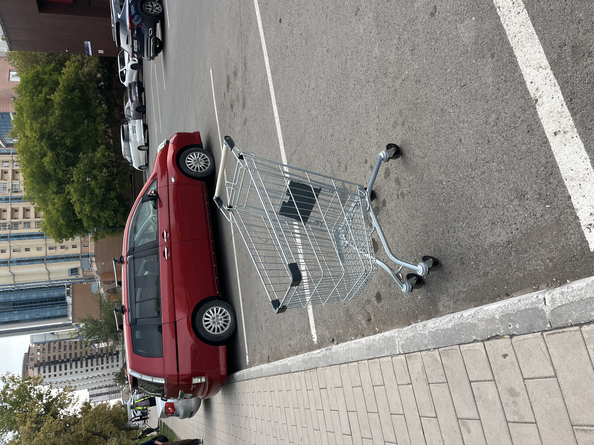 In a parking lot in Astana, Kazakhstan, a red minivan is parked adjacent to a sidewalk. A shopping cart is positioned in the parking lot, near the curb. Several other vehicles, including a white SUV and a dark blue SUV, are parked in the background. The scene appears to be in an urban environment with buildings and trees visible. The weather is overcast.