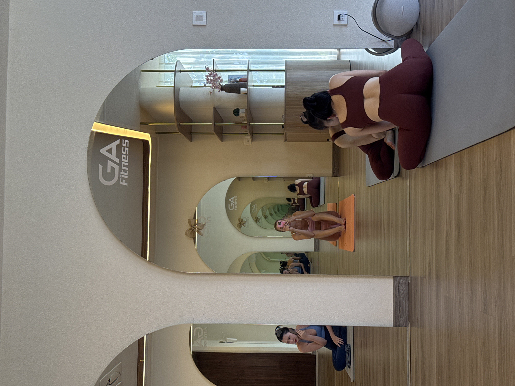 Here is a description of the image:
The image shows the interior of a yoga studio, likely in Unknown, Unknown, as indicated by the "GA Fitness" logo visible on a mirror. A woman is kneeling on a gray yoga mat, wearing a maroon athletic top and leggings. Her back is to the camera. The room contains multiple arched mirrors, creating a tunnel effect and reflecting other individuals in yoga poses. One woman, wearing a light-colored athletic top and bottoms, is sitting in a cross-legged position on an orange yoga mat, looking at her phone. Another woman, in blue athletic wear, is also visible, likely in a similar pose. The walls are a light color, and the floor is a wood laminate. There are shelves on the wall behind the kneeling woman, and various objects, including what appears to be an air purifier, are also present. The overall impression is of a clean and modern fitness studio with multiple participants.
