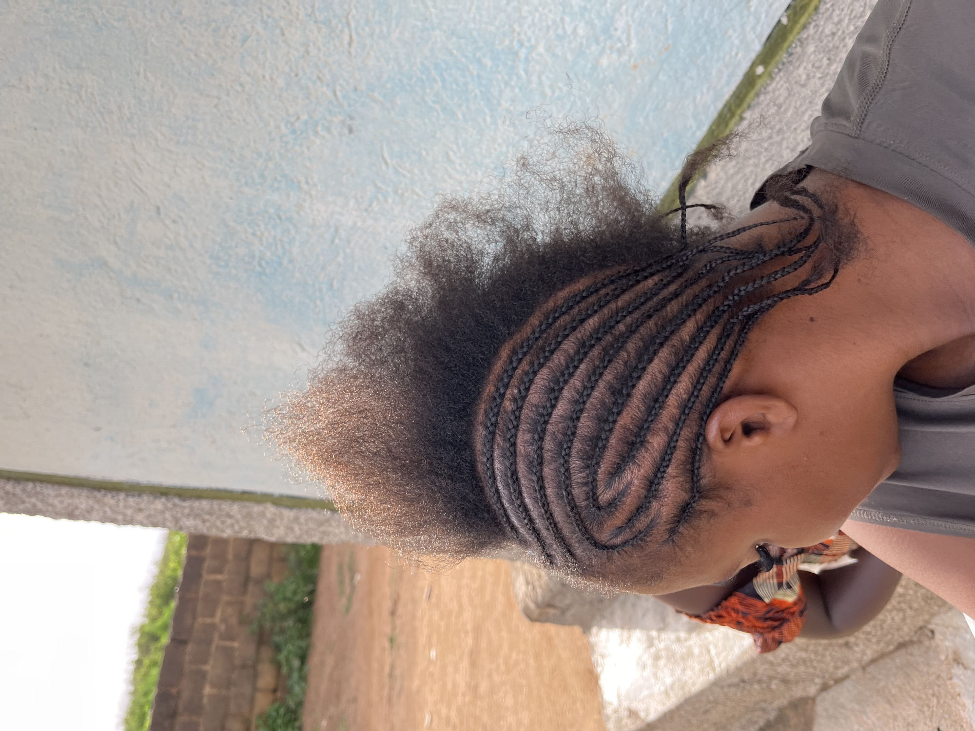 A young Black person is having their hair braided in a cornrow style. The person is seen from the side, with their head tilted downwards. Their natural hair is dark brown and appears to be thick and curly. The braids are neatly done, starting from the front of their head and curving towards the back. Some of the natural hair at the crown of the head is left loose and appears fluffy. The person is wearing a grey t-shirt, and a colorful scarf or fabric is visible around their neck or chest area.
The background is somewhat out of focus. The immediate surface below the person appears to be made of light-colored stone or concrete. Behind that, there is a wall constructed of reddish-brown bricks, with some green foliage growing on or near it. Further in the background, a bright white area suggests an open space, possibly outdoors with bright sunlight. A textured wall with patches of blue and white is visible on the upper left side, suggesting a building or outdoor structure. The lighting suggests it is daytime, likely a sunny day, given the brightness of the white area. The setting appears to be outdoors in a casual, possibly residential or communal area. No text is visible in the image.