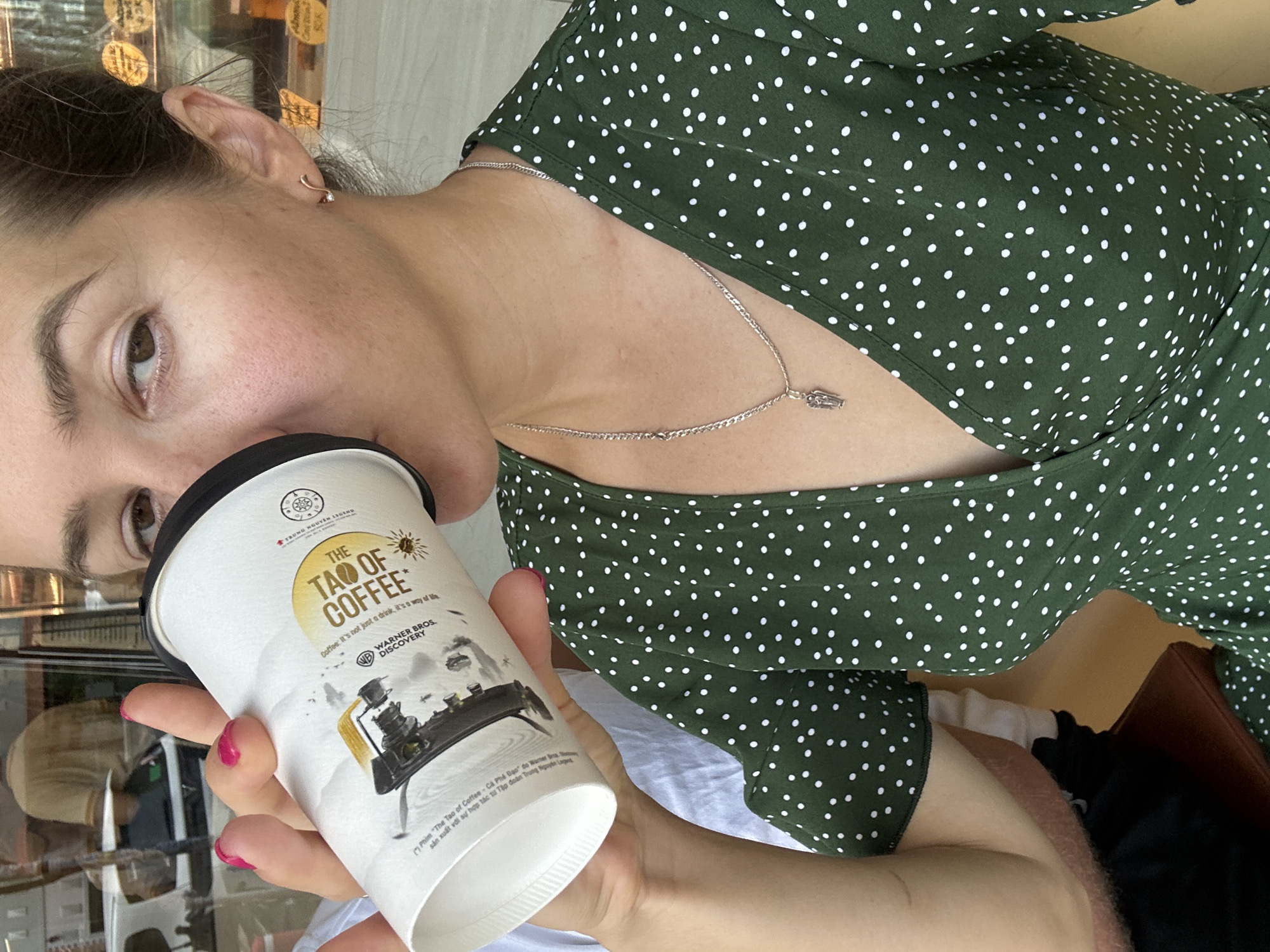 A medium shot shows a woman drinking from a paper cup of coffee. She is wearing a green polka dot dress and a silver necklace with a small pendant. In the background, a blurred reflection shows a street scene in Nha Trang Ward, Vietnam, with people walking and shops lining the street. The woman's hand, painted with bright pink nail polish, is holding the cup to her lips. The coffee cup has branding that reads "THE TAO OF COFFEE." The overall lighting suggests it is daytime.
