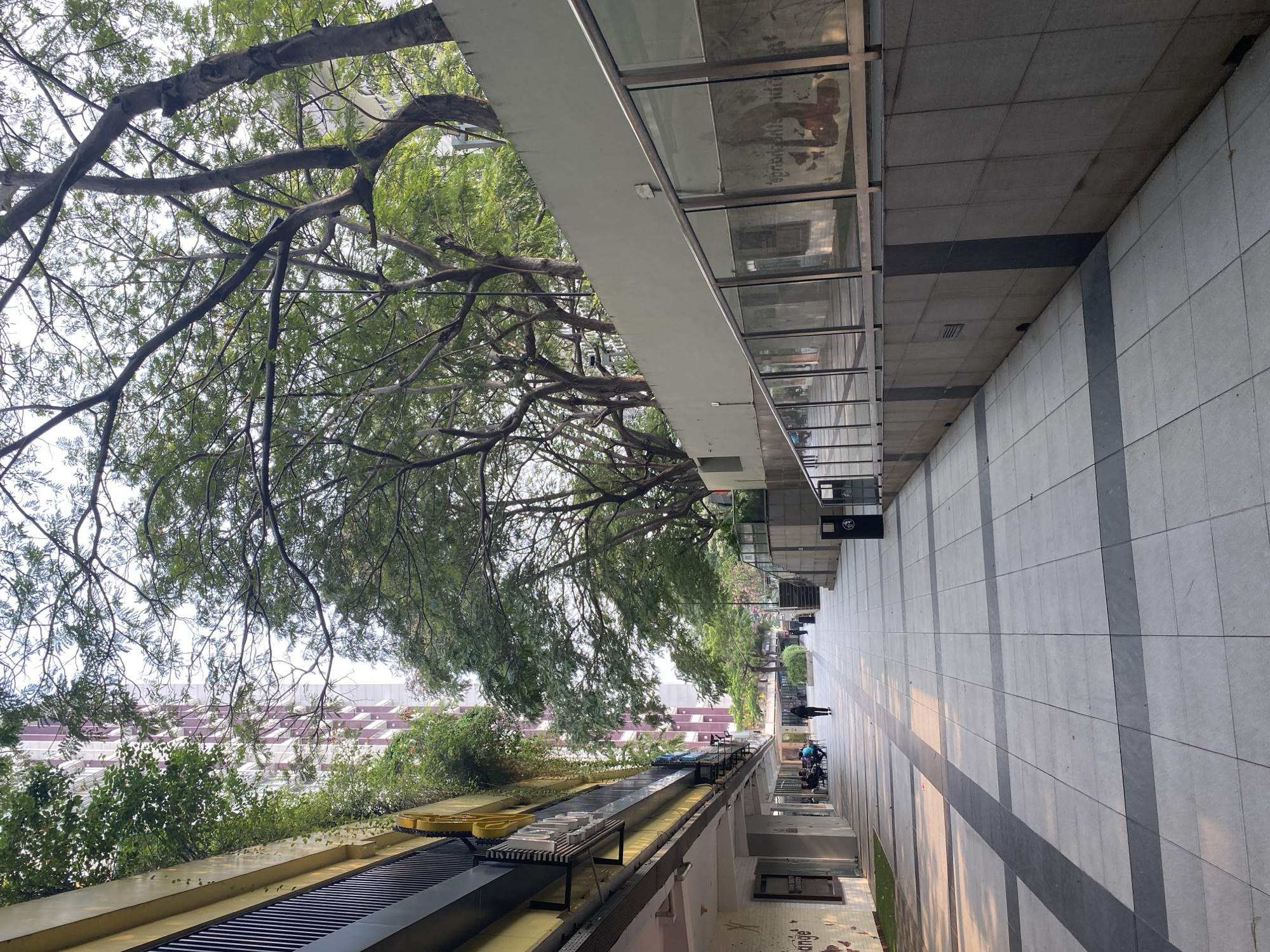 The image depicts a sun-drenched pedestrian walkway lined with shops on the left and a raised area with greenery and modern architecture on the right. The walkway is paved with large square tiles, alternating in color with darker rectangular strips. In the distance, a few individuals are visible walking. On the left, a building with several floors is partially obscured by lush green vegetation. The right side of the walkway features a grey concrete wall topped with a railing and glass panels, behind which stairs ascend. The scene suggests an urban environment, potentially a shopping district or public space within a city, with a mix of commercial and green spaces. Based on its architectural style and the presence of tropical trees, this location is identified as Baywalk Mall, located in North Jakarta, Indonesia.