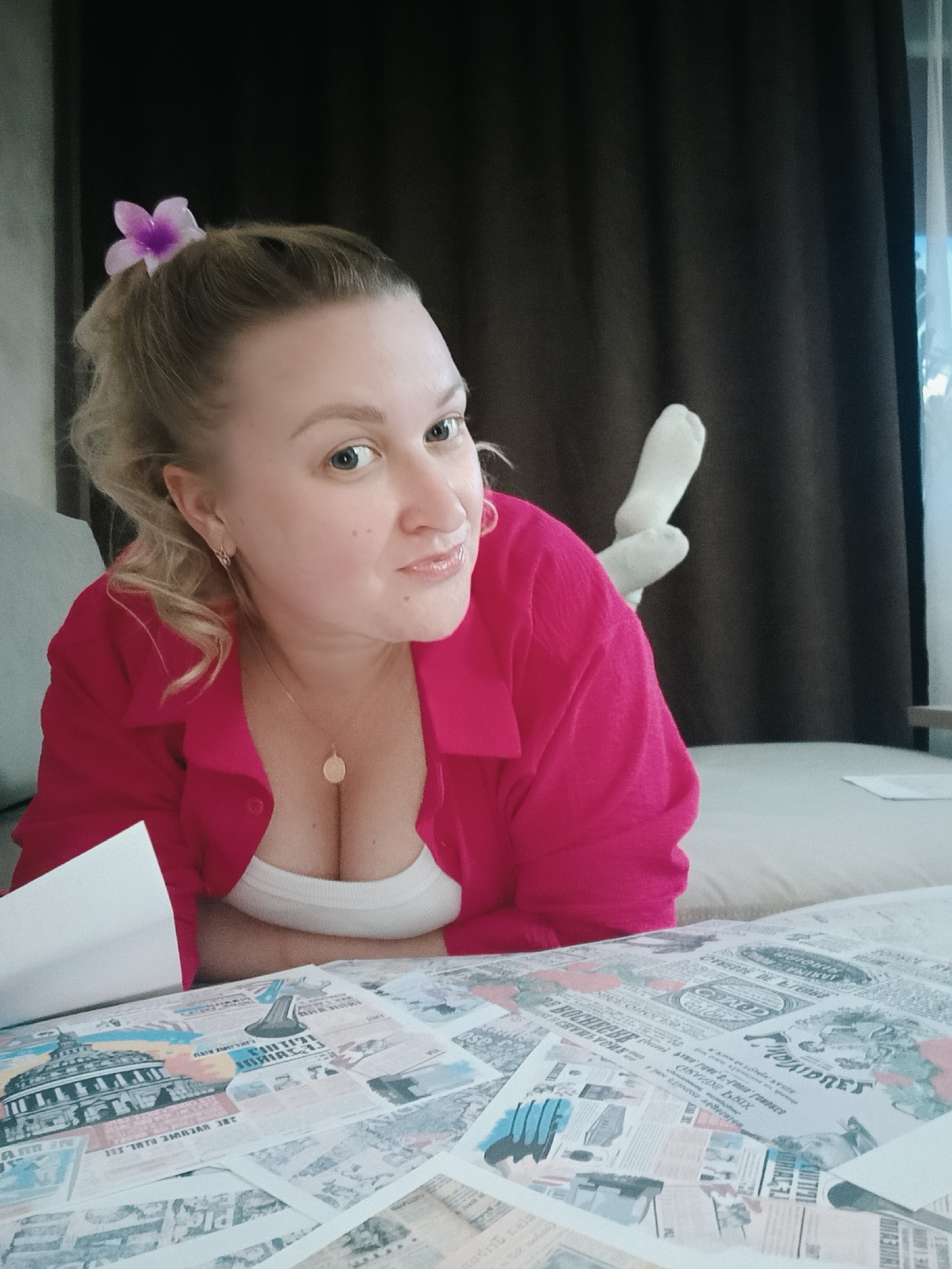 A woman is depicted indoors, likely in a bedroom or lounge area, lying on her stomach or side, propped up on her elbows. She has light-colored hair, styled partially up, with a small purple flower tucked into it. Her gaze is directed at the camera, accompanied by a subtle smile. She wears a vibrant pink collared shirt, unbuttoned over a white camisole, revealing a gold-colored circular pendant necklace. A gold hoop earring adorns her right ear. In the background, her feet, clad in white socks, are visible, and behind them, dark brown curtains and a window offering natural light are present. The foreground features a surface covered with an assortment of papers, resembling vintage newspaper clippings or decorative prints, displaying various texts, possibly in Cyrillic, and illustrations, including one of a building reminiscent of the US Capitol. The overall scene conveys a relaxed and casual atmosphere.