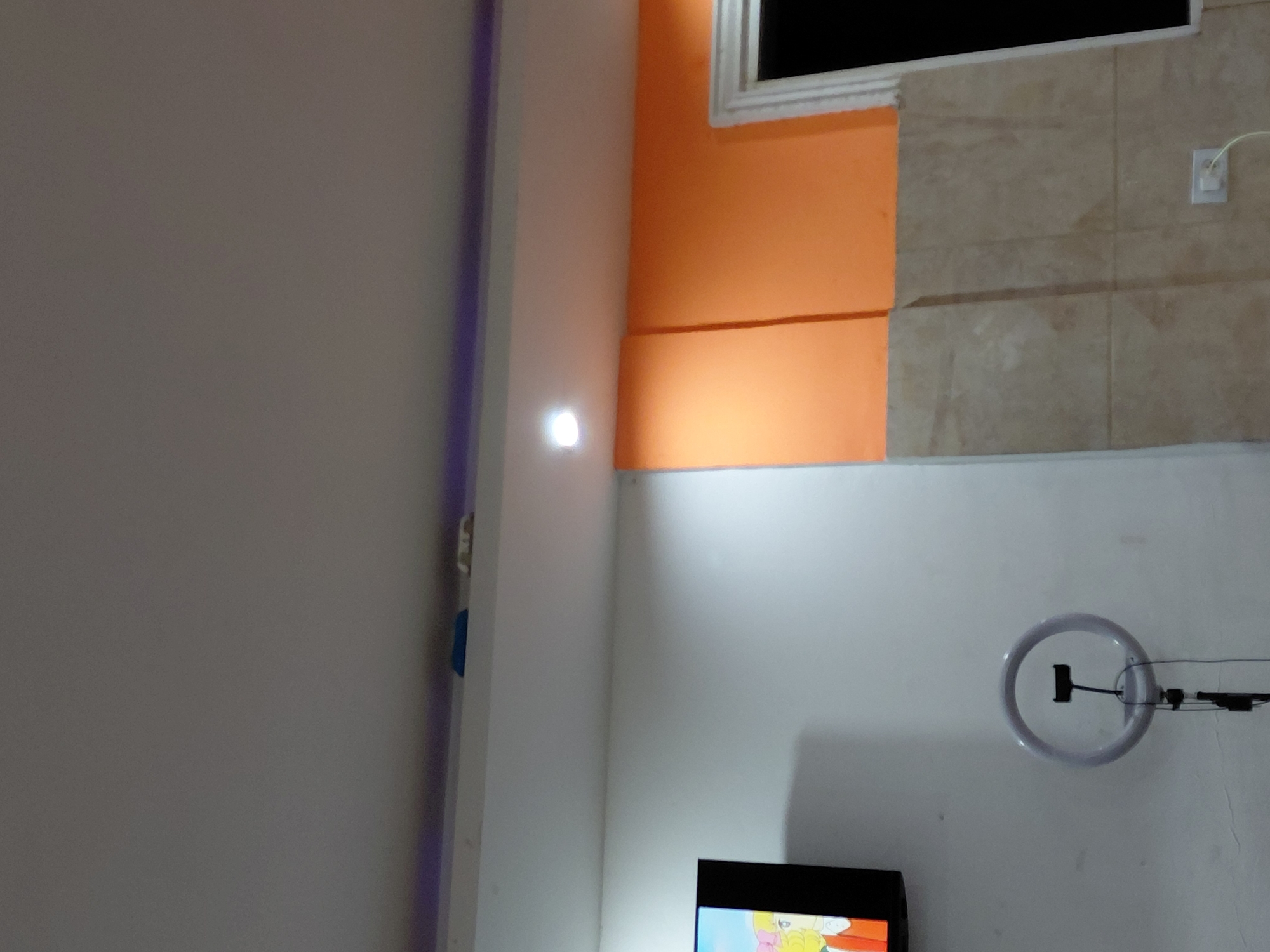 A low-angle interior shot depicts a corner of a room with multiple wall surfaces. The left wall is white, meeting a perpendicular white wall adjacent to it that is adorned with a vertically oriented LED strip in the upper left. A light fixture is mounted on the wall. An orange section of wall is visible in the upper right, bordered by an off-white section above. A square section of tan and white tiled wall is present. A white electrical outlet is mounted on the tiled wall. On the right side of the image, a white wall displays a ring light mounted on the wall. At the bottom of the image, a black television screen is partially visible, displaying cartoons. The location is purportedly Santa Ana, Venezuela.