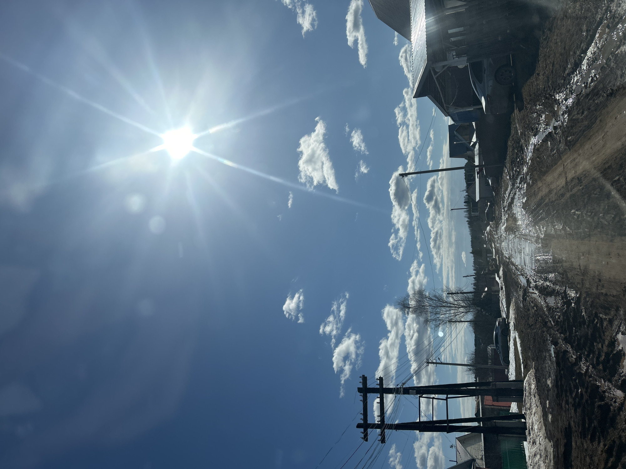 The image depicts a rural street in Фоки, Russia on a bright, sunny day. A dirt road, partially covered in mud and melting snow, runs along the right side of the frame. Buildings, including houses and a shed-like structure, line the street. Power lines and utility poles are visible, cutting across the sky. The sky is blue with scattered white clouds. The sun is positioned high in the sky, creating a bright glare.