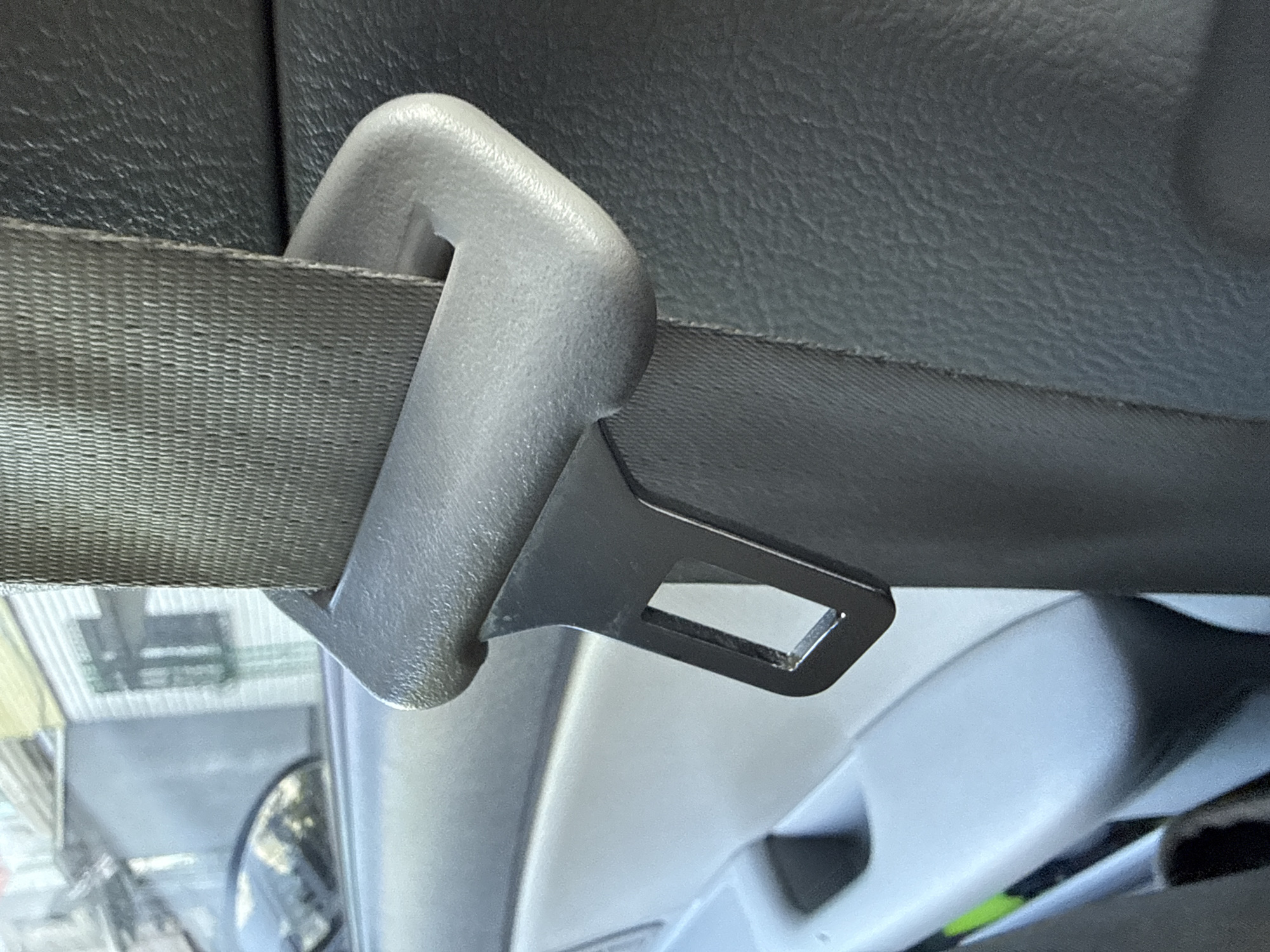 The image shows a close-up, angled shot of a car's interior, focusing on a gray seatbelt buckle mechanism. The seatbelt webbing, with its textured pattern, is visible entering the buckle. The buckle itself is a two-part system: a light gray plastic housing that appears to be attached to the car's interior trim, and a black metal latch plate with a rectangular opening. This metal latch plate is attached to the seatbelt and is designed to slide into the gray housing to secure the belt.
The background of the image reveals more of the car's interior. To the left, there is a dark, textured surface, likely part of the car's upholstery or trim. Above and to the right of the seatbelt mechanism, a lighter gray, smooth surface is visible, possibly a headliner or another interior panel. In the lower portion of the image, out of focus, a reflection is visible on a shiny surface, showing parts of what appears to be the car's exterior, including windows with some objects or structures outside. The overall lighting suggests it is daytime, possibly indoors or in a shaded area, given the soft light and lack of harsh shadows. There is no visible text in the image. The scene is static, with no discernible activity other than the depiction of a secured seatbelt mechanism.