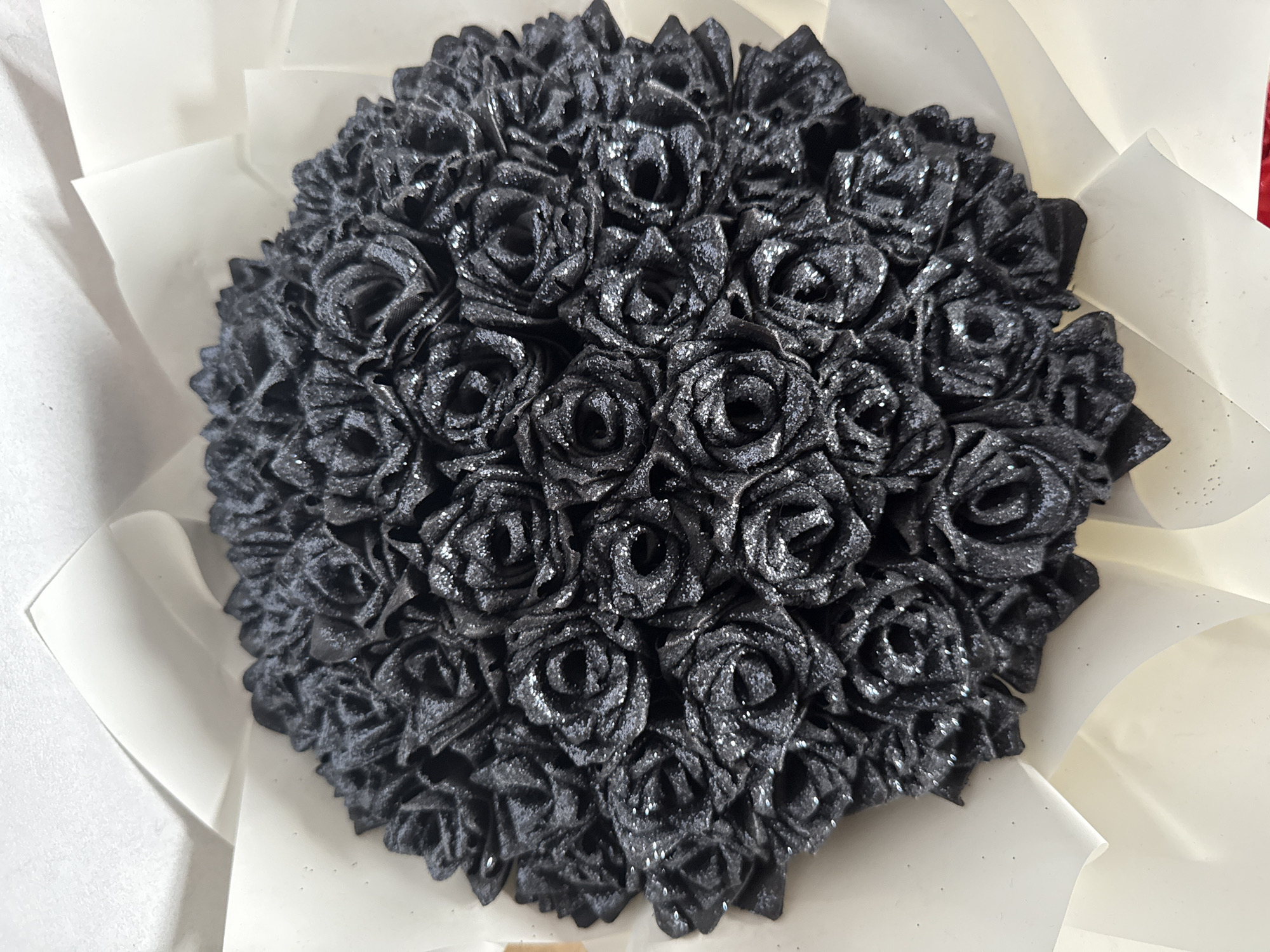 A close-up shot of a bouquet of black, sparkly roses arranged in a dense, circular formation. The roses are surrounded by a white paper wrap. There is no visible information indicating the location is Votkinsk, Russia. The environment is plain and undetailed, focusing primarily on the bouquet. There are no people or interactions visible in the scene.