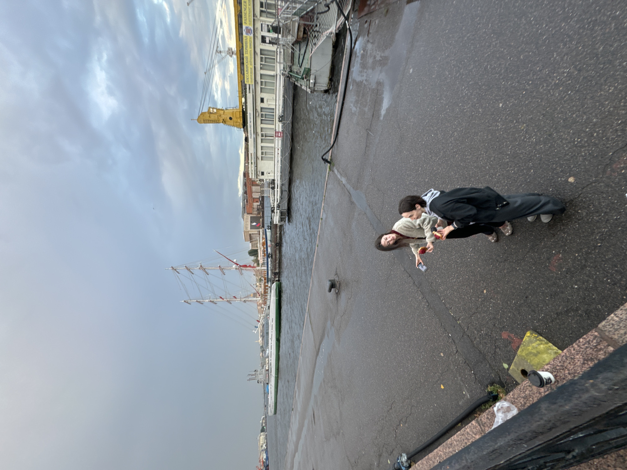 Two individuals are standing on a wet asphalt promenade next to a body of water. One individual, on the left, is wearing a light-colored hooded jacket and black pants, holding a red object in their right hand. The other individual, on the right, is wearing a dark-colored hooded jacket and gray pants, also holding a red object in their right hand. In the background, a large sailboat with multiple masts and sails is docked. To the right of the sailboat is a long, white building with windows, identified by Russian text as a passenger terminal. A yellow crane is visible above the terminal. The sky is overcast with gray clouds. The scene appears to be a waterfront area in Saint Petersburg, Russia, likely a port or harbor, given the presence of the large sailboat, terminal building, and crane.