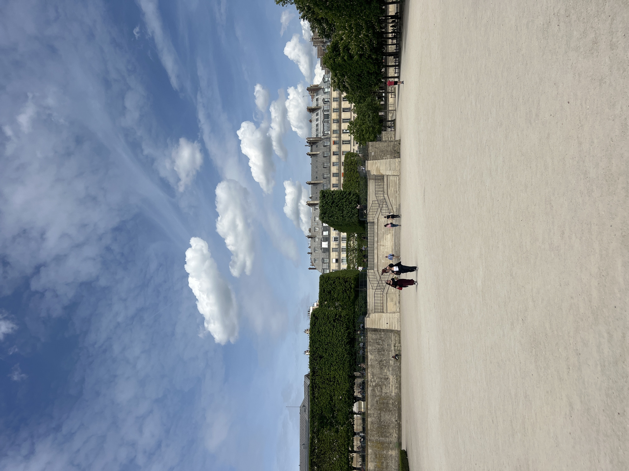 This daytime photograph captures a wide outdoor scene in a Parisian park or garden. The foreground and much of the middle ground are dominated by a vast, open expanse covered in light-colored sand or gravel. Along the left side of this open area, a neatly trimmed line of green trees runs parallel to a low stone wall, behind which additional trees and sections of classic multi-story buildings are visible. These buildings feature typical Parisian architecture with light facades, numerous windows, and grey mansard roofs.
Several individuals are present in the scene. In the mid-ground on the left, a set of stone steps ascends, and a small group of people can be seen near its base, some standing and others possibly walking. Further in the distance, more small figures are scattered across the sandy ground and near the structures.
The upper portion of the image is filled with a bright blue sky, adorned with a variety of white clouds. These include distinct, fluffy cumulus clouds, particularly concentrated above the buildings, and also some thinner, wispy cirrus clouds stretching across the sky. The clear lighting suggests a pleasant, sunny day, likely during the late morning or afternoon. The overall atmosphere is calm and serene, depicting an everyday moment in a public green space in Paris, France. No discernible text is visible in the image.