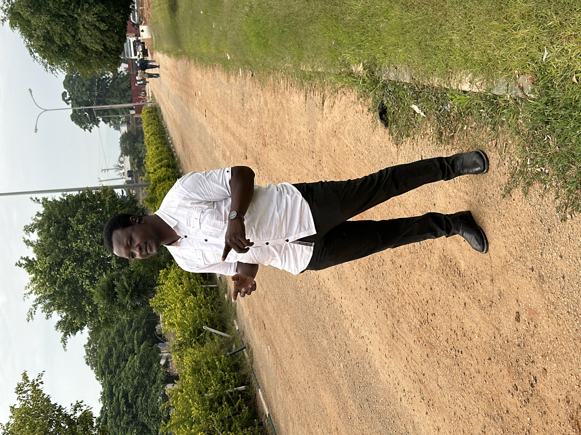 A man in a white collared shirt and black pants stands on a dirt road in Dadi Riba, Nigeria. He is wearing black earbuds and a watch on his left wrist. His right hand is raised with his index finger extended, and his left hand is in a similar gesture. He is looking to his right. The road is flanked by green foliage and trees. In the background, several people are walking, and a white truck is parked. A street lamp is visible on the right side of the road. The sky is overcast.