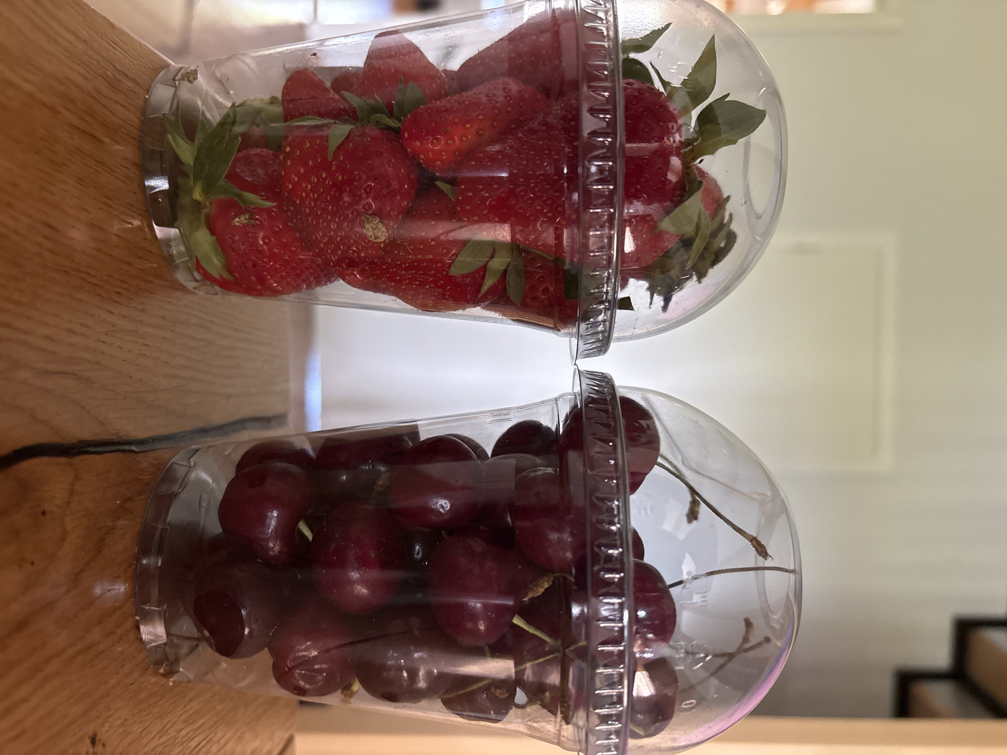 Two clear plastic cups are stacked vertically on a wooden surface. The top cup contains strawberries with green stems and leaves visible. The bottom cup contains cherries with stems visible. The background is blurred and shows a light-colored wall and a glimpse of what appears to be a staircase. The location in Chaikovsky, Russia is not known and could not be identified.