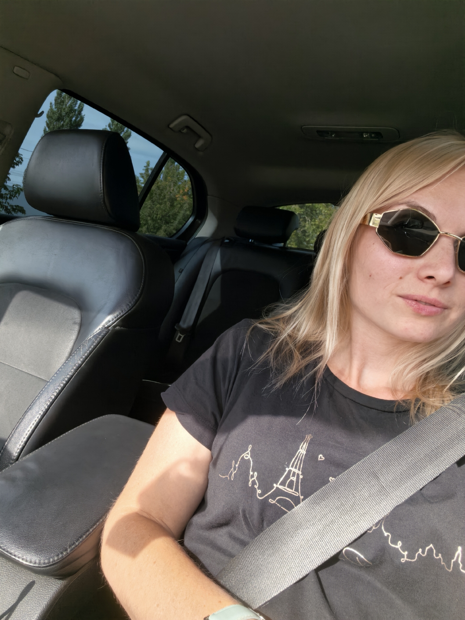 A blonde woman with sunglasses sits in the passenger seat of a car, wearing a black t-shirt with a line drawing of the Eiffel Tower. The car interior is visible, including black leather seats and a seatbelt. Outside the window, trees and a blue sky can be seen. The scene appears to be a daytime shot, likely taken in Sterlitamak, Russia.