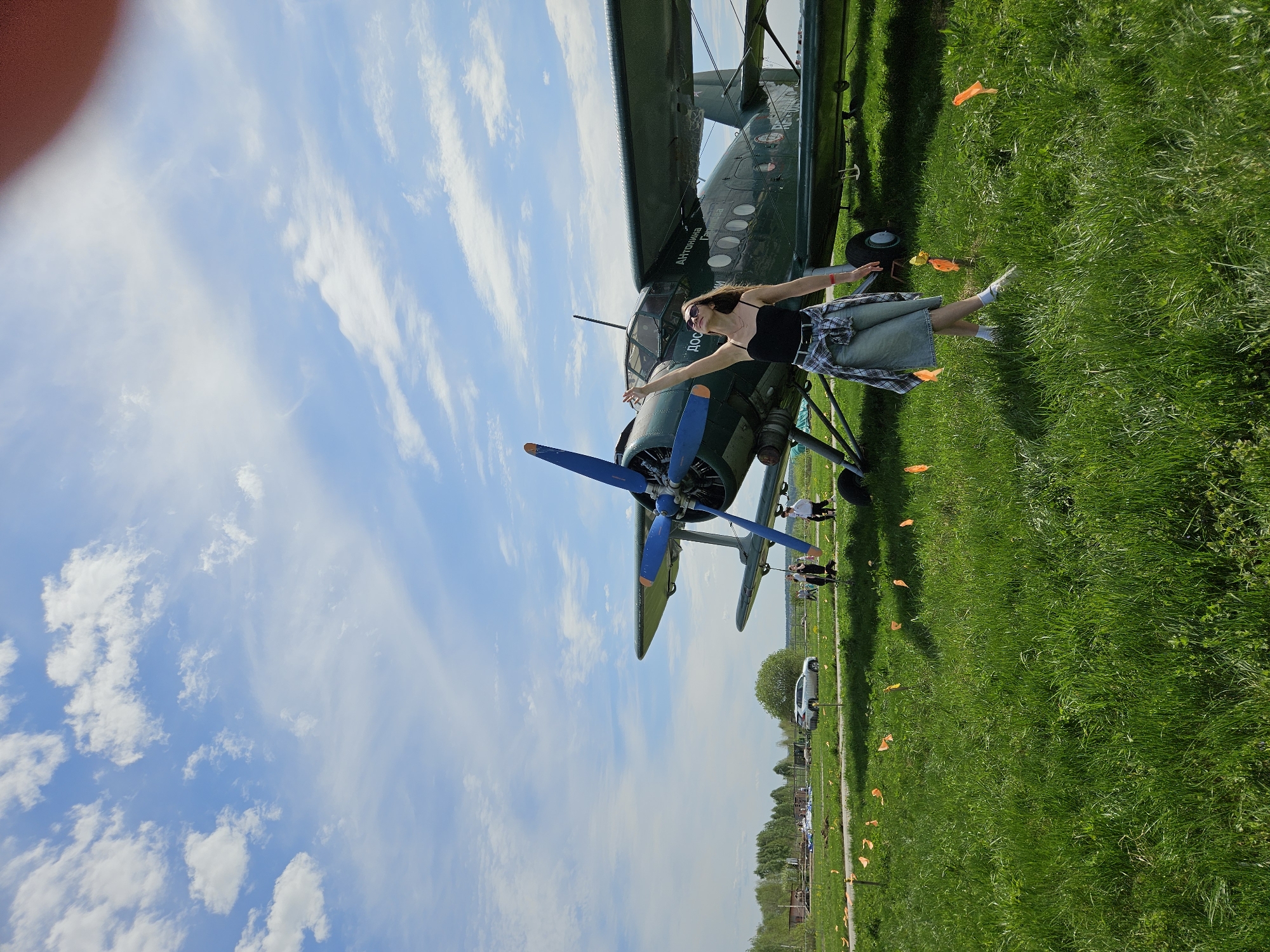 A young woman stands in front of a green biplane, arms outstretched, in Izhevsk, Russia. The sky is blue with scattered clouds. The woman wears a black tank top, denim skirt, a plaid shirt tied around her waist, white socks, and white sneakers. She is also wearing sunglasses. The biplane is green with blue propellers. The ground is covered in green grass with orange flags scattered about. In the background, a white car is parked near trees. Other people are visible behind the biplane.
