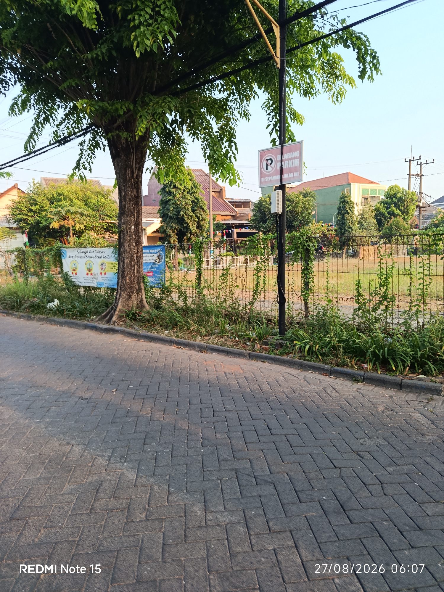 The image depicts a street scene in Keboansikep, Indonesia. A paved road with a herringbone pattern made of dark gray bricks occupies the foreground. To the right of the road, a metal fence encloses a grassy area with various plants and vines growing on and around it. Beyond the fence, several houses with reddish-brown and green roofs are visible, interspersed with trees and utility poles. A large tree with dark bark and green foliage stands prominently on the left side of the image, partially obscuring the view of the houses. A sign with a red circle and a diagonal line over a "P" symbol, indicating "DILARANG PARKIR" (No Parking), is mounted on a pole in front of the fence. Two banners are also attached to the fence; one is white with colorful illustrations and text that appears to be in Indonesian, and the other is blue with text that is partially obscured. The sky is clear and bright blue, suggesting a sunny day. The bottom left corner of the image displays "REDMI Note 15" and the bottom right corner shows "27/08/2026 06:07". There are no people visible in the scene.