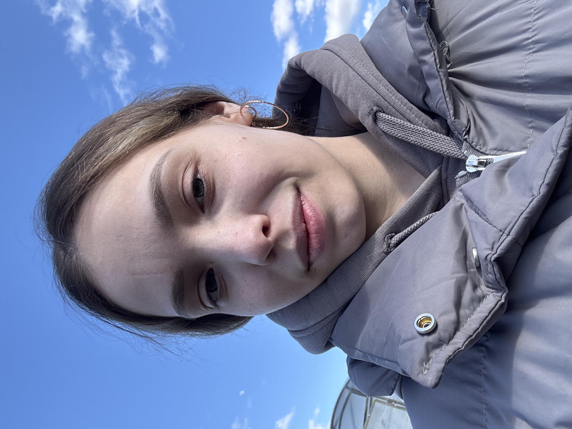 A young woman is taking a selfie outside against a clear blue sky with scattered white clouds. She is wearing a gray hooded sweatshirt and a gray puffy jacket. She has light brown hair and is wearing hoop earrings. A partial view of a greenhouse is visible in the lower right corner. The location is likely in Кемуль, Russia.