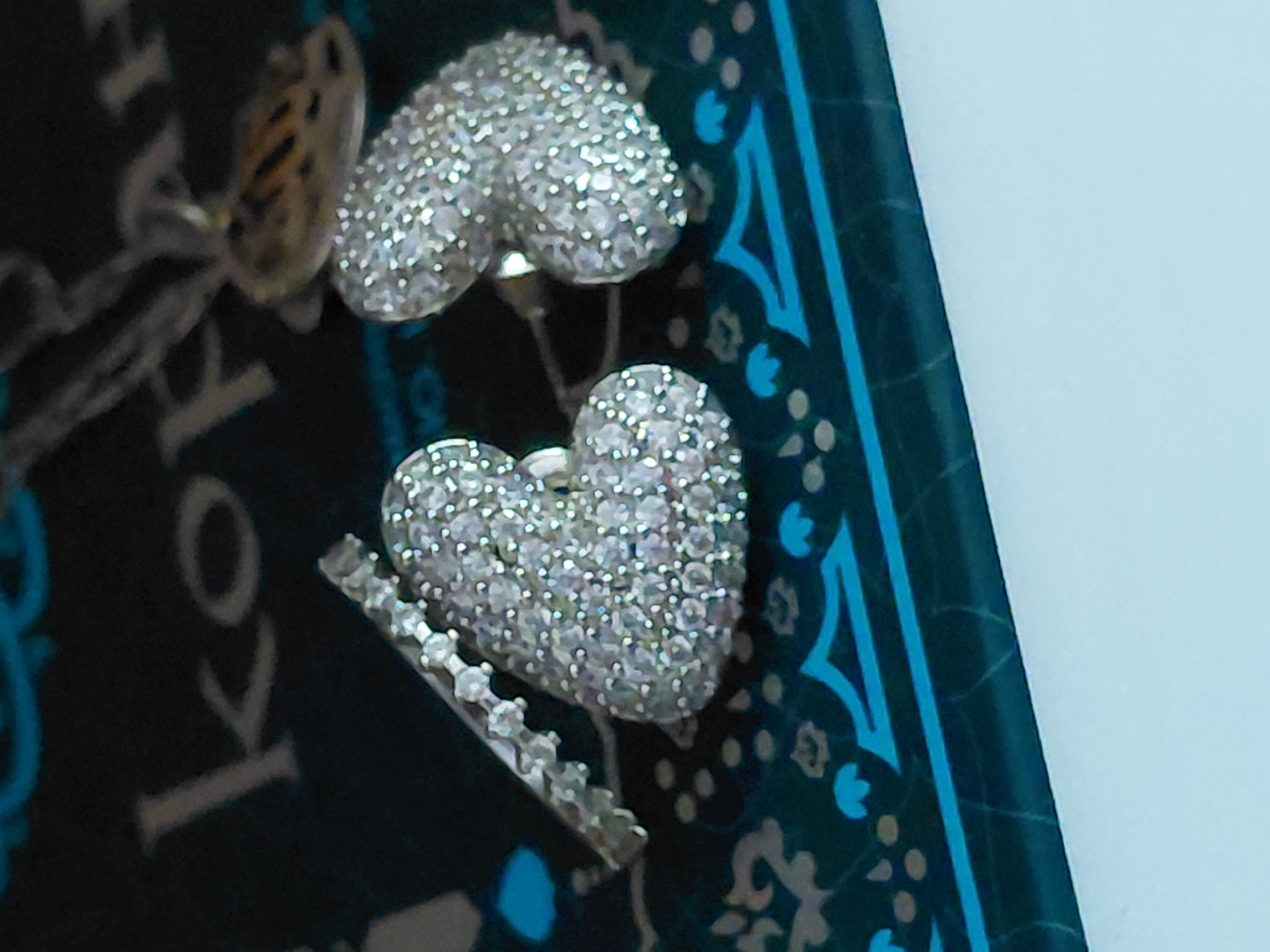 A pair of heart-shaped earrings encrusted with small, clear gemstones are displayed on a dark, patterned surface. The earrings are a bright, metallic silver color, and they are positioned slightly apart from each other. To the left of the earrings, a thin, jeweled band is visible, which appears to be part of a ring. The surface they rest on has a dark blue-green hue and features intricate, lighter blue and gold patterns. In the background, some blurry text is visible, reading "KORHAN". The overall scene suggests a close-up product shot or a display of jewelry. There is no indication of the city of Novosibirsk, Russia.