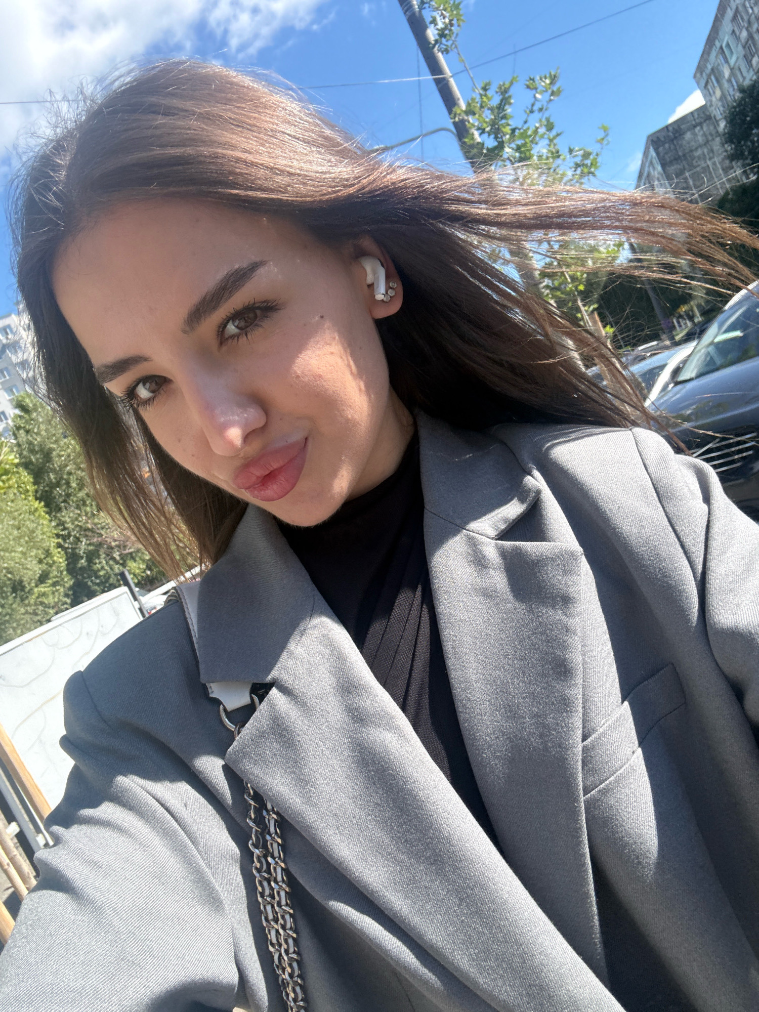The image is a selfie taken outdoors in the city of Chișinău, Moldova. A young woman with long, brown hair and light skin is the primary subject. She is wearing a gray blazer over a black top and white wireless earbuds. The background includes a bright blue sky with some clouds, green trees, and buildings. A vehicle is partially visible in the background. The lighting suggests it is daytime, likely sunny.