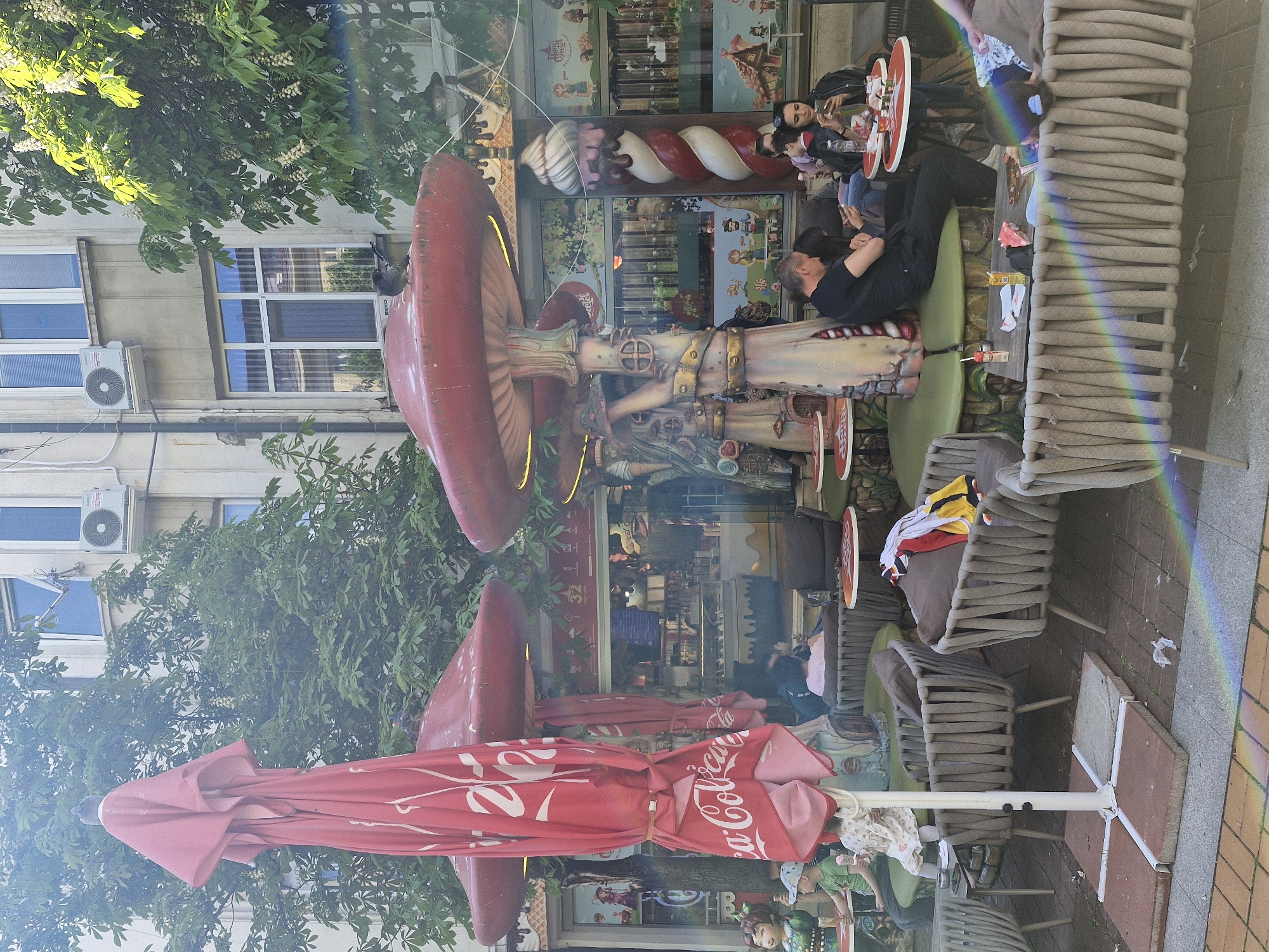 The image captures an outdoor scene in Sofia, Bulgaria, showcasing a whimsical cafe. The cafe exterior is adorned with large decorative mushrooms and candy cane-like pillars. People are seated at tables, some looking at their phones while others are seated with young children. In front of the cafe is an outdoor terrace with tables and chairs under a closed Coca-Cola branded umbrella. The street has a sidewalk with chairs and some clothing sitting on the chairs. A building is partially visible in the background with windows and AC units. Trees and greenery are also present, adding to the outdoor ambiance.