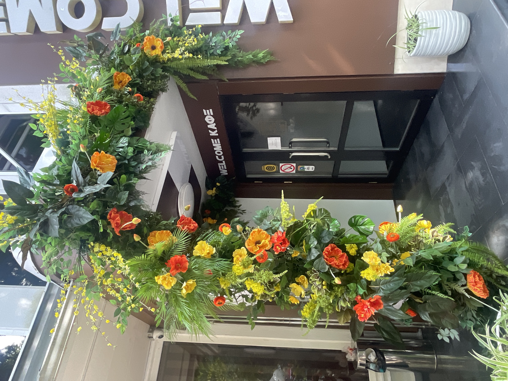The image shows a storefront decorated with a large floral archway made of lush green foliage and vibrant orange and yellow flowers. The arch spans across the entrance of a building. Above the double glass doors, the words "WELCOME KAФE" are displayed in white, block lettering on a brown facade. To the right of the entrance, large white letters spelling "COMC" are affixed to the building. The entrance itself features dark-tinted glass doors with silver handles. A small potted plant with thin green leaves sits on the tiled step to the right of the door. The overall setting suggests an outdoor cafe entrance in an urban environment. The signage indicates the location is likely Sochi, Russia.