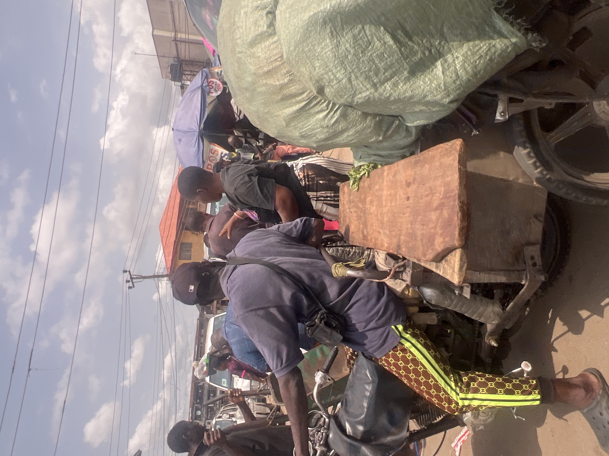 This image captures a busy street scene in Onitsha, Nigeria. Numerous people are clustered together, suggesting a marketplace or a transportation hub. Individuals are riding motorcycles loaded with goods, including a large sack covering nearly half of the upper portion of the frame. A man wearing patterned pants sits on one motorcycle, while others are gathered around, possibly engaging in commerce or waiting for transportation. The environment is dense, with buildings and power lines visible in the background. The light suggests it is daytime, possibly midday.