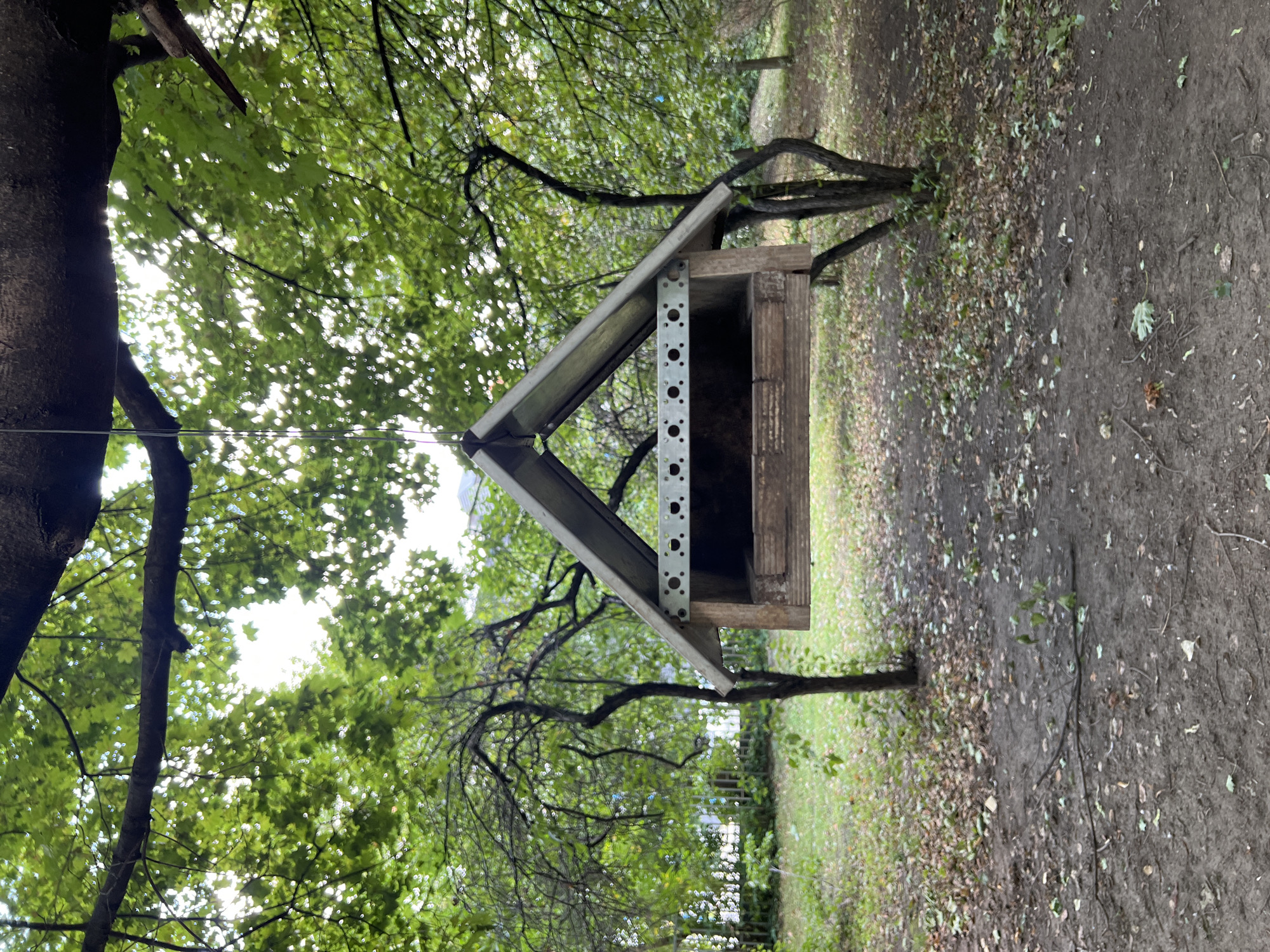 The image depicts a wooden bird feeder with a metal roof, suspended from a tree branch. The feeder is located in an outdoor setting with lush green trees and foliage. The ground beneath the feeder is covered with dirt and fallen leaves, indicating an autumnal or post-autumnal season. The overall atmosphere is serene and natural. The background suggests an urban environment, with hints of buildings visible through the trees, possibly in the city of Moscow, Russia. There are no people or animals visible in the frame, and no apparent actions or interactions are taking place. The focus is on the bird feeder and its natural surroundings.