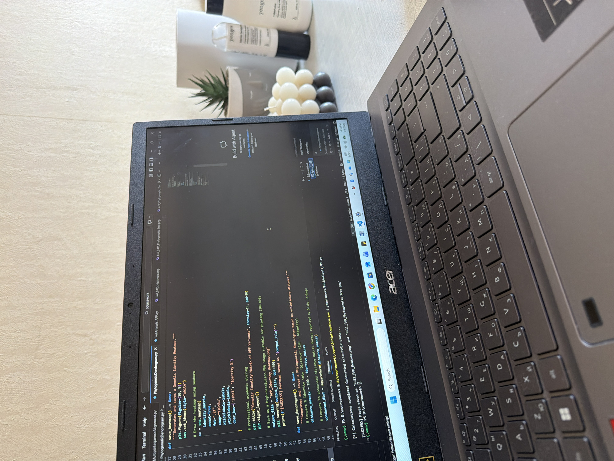 The image shows a laptop displaying Python code related to phylogenetic analysis and heatmap generation. The laptop is an Acer brand and the keyboard has Cyrillic characters, suggesting the user is located in a region where this script is used. Adjacent to the laptop are a few decorative items: a small succulent in a grey pot, a decorative candle made of white and grey wax spheres, and several white bottles with black labeling from "yenges". Given the Cyrillic keyboard, the location is potentially Sofia, Bulgaria. The laptop is currently displaying the output from running the Python script, with messages indicating successful creation and saving of heatmap and phylogenetic tree plots. The code itself appears to be related to processing genetic identity data. The overall scene depicts a workspace with a computer and some decorative items.