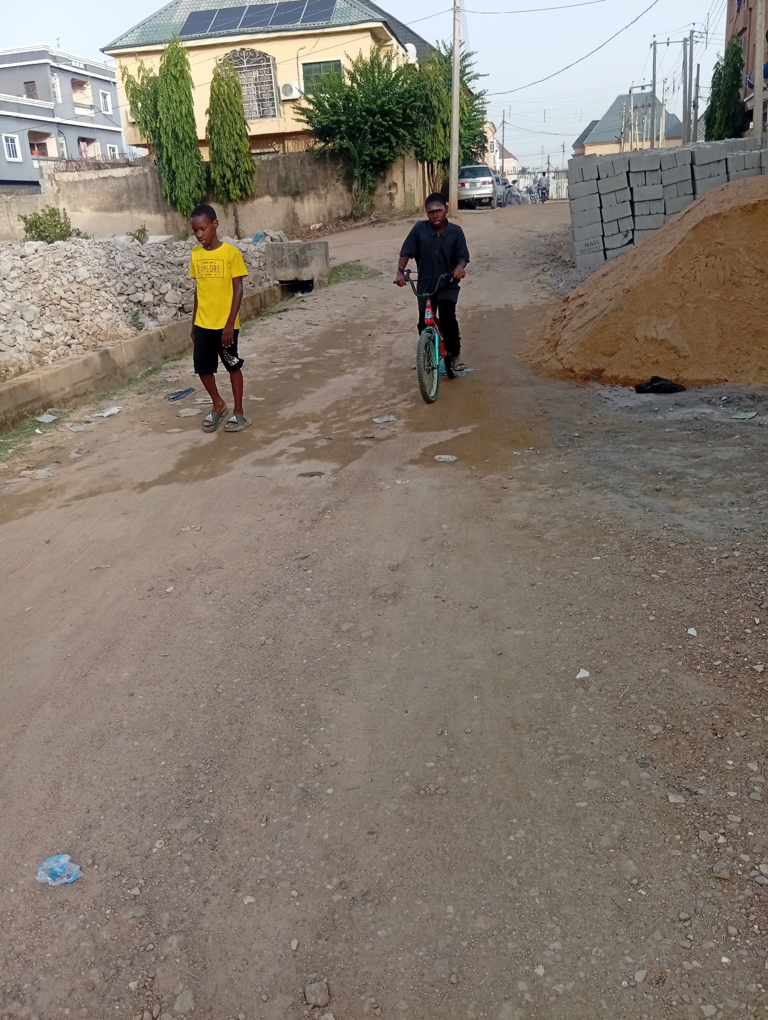 The image depicts a dirt road in Aviation Quarters, Nigeria. A boy in a yellow t-shirt and black shorts walks along the left side of the road, while another boy rides a bicycle on the right. A pile of rubble is on the left side of the road. A mound of sand and a stack of cement blocks are to the right. In the background are buildings, cars, and power lines. A plastic bag lies on the ground in the foreground.