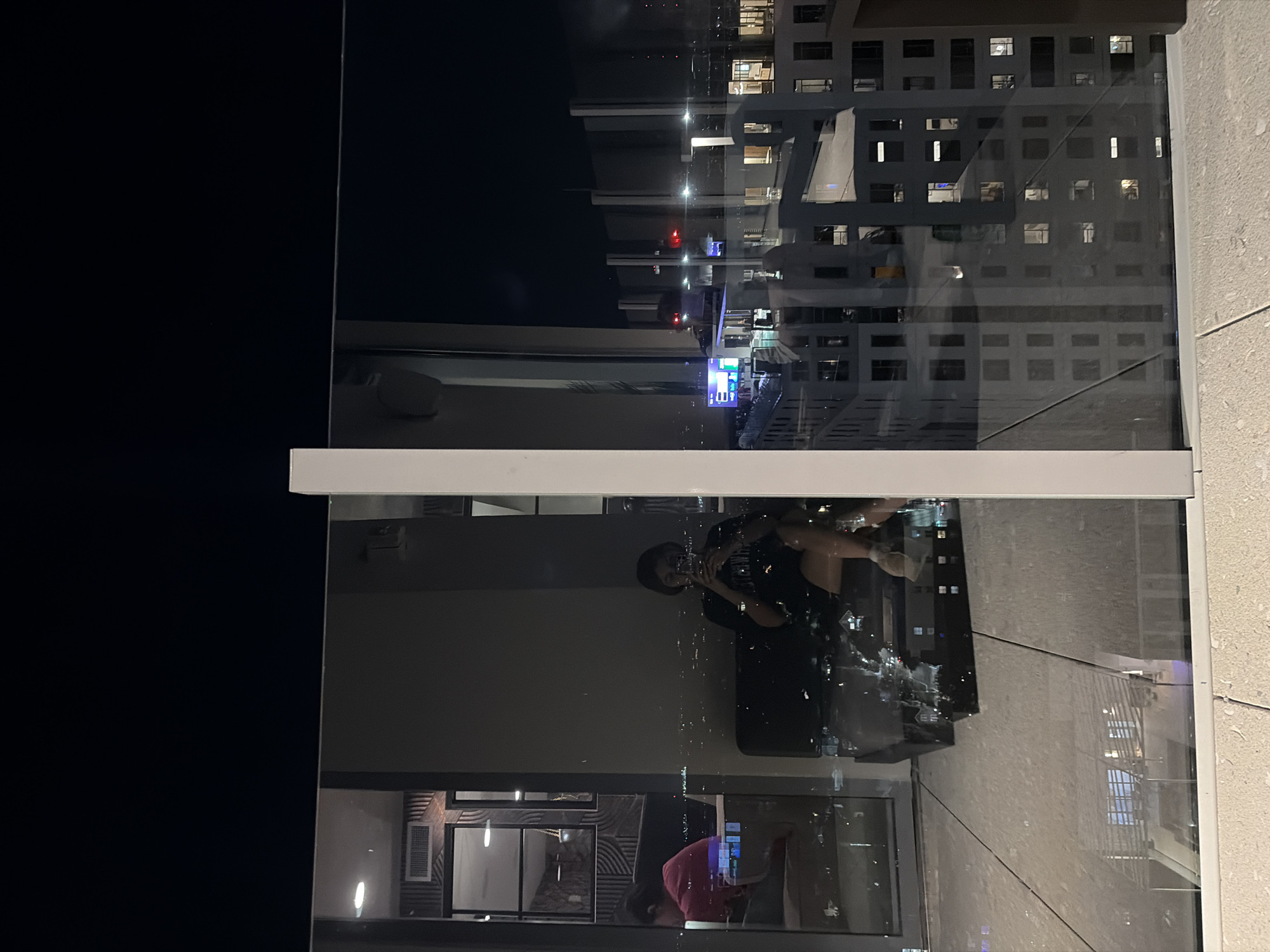 A nighttime indoor scene appears to be taken from a high-rise balcony or window in Austin, United States of America. The view through the glass features a reflection of a person sitting on a couch or chair inside the room. The person is wearing a black t-shirt. The reflection also shows a portion of the interior room. Outside the glass, one can see a cityscape at night with illuminated buildings.