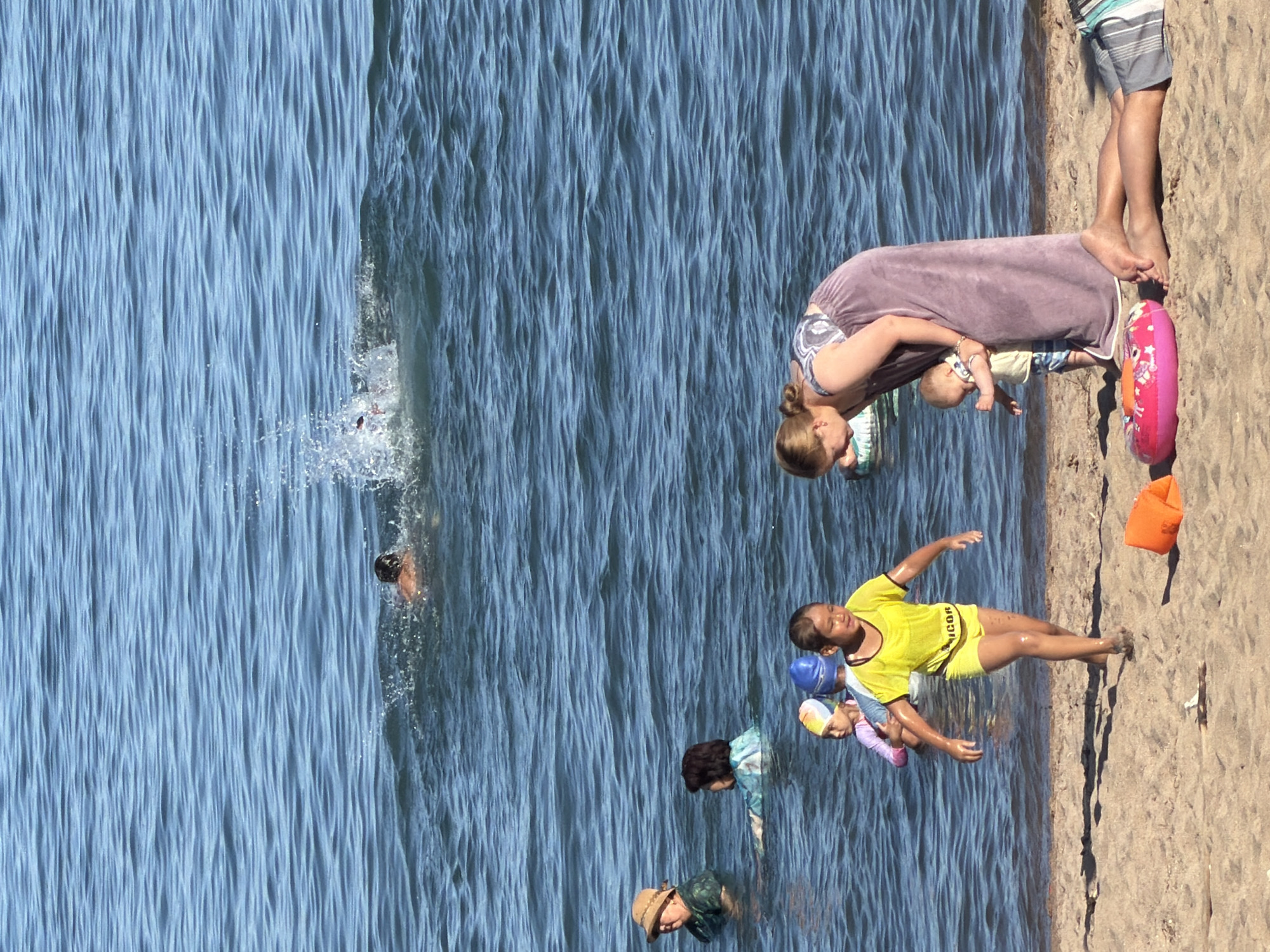 The image depicts a sunny day at a beach where several people are enjoying the water. In the foreground, on the sandy shore, a young child in a yellow swimsuit runs towards the water. Beside the child, a woman in a long, purple cover-up holds a baby. Nearby, an orange float and a pink inflatable ring are on the sand. In the water, several other individuals are visible. Two children in swim caps are wading, and further out, adults are swimming or standing in the water. In the background, a person is swimming and creating a splash. The water is a clear blue with gentle ripples, and the sky is not visible in the frame.