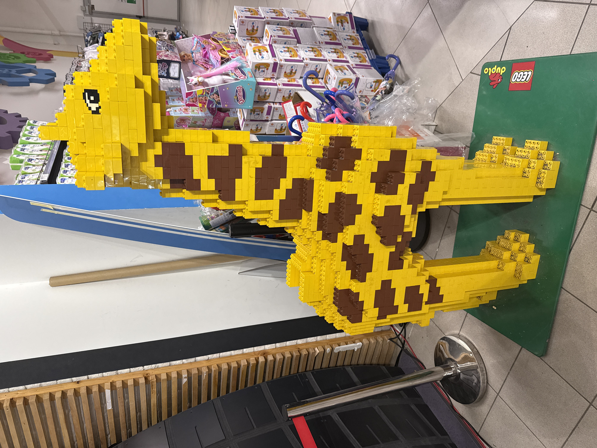 A large, intricately constructed giraffe model, fashioned from yellow and brown LEGO Duplo bricks, is prominently displayed indoors, likely within a toy store or a supermarket's toy section in Zagreb, Croatia. The giraffe features a distinct spotted pattern across its body and tail, with pixelated black and white bricks forming its eyes on the yellow head.
The model is positioned above a black partition or barrier, with a chrome stanchion partially visible in the foreground, suggesting a designated display area. To the right, the giraffe rests on a large green mat prominently featuring the red "LEGO" logo and white "Duplo" text, confirming the brand.
The background reveals shelves and stacks of various products, including numerous cardboard boxes, toys like a unicorn and a doll, and other merchandise. The brand name "Dreamzzz" is visible on some blue product boxes. Blue and red plastic items, possibly toy accessories or handles, are also scattered behind the display. The floor consists of light-colored tiles, and the scene is brightly illuminated by artificial lighting, characteristic of a retail environment. No people are visible in the image.