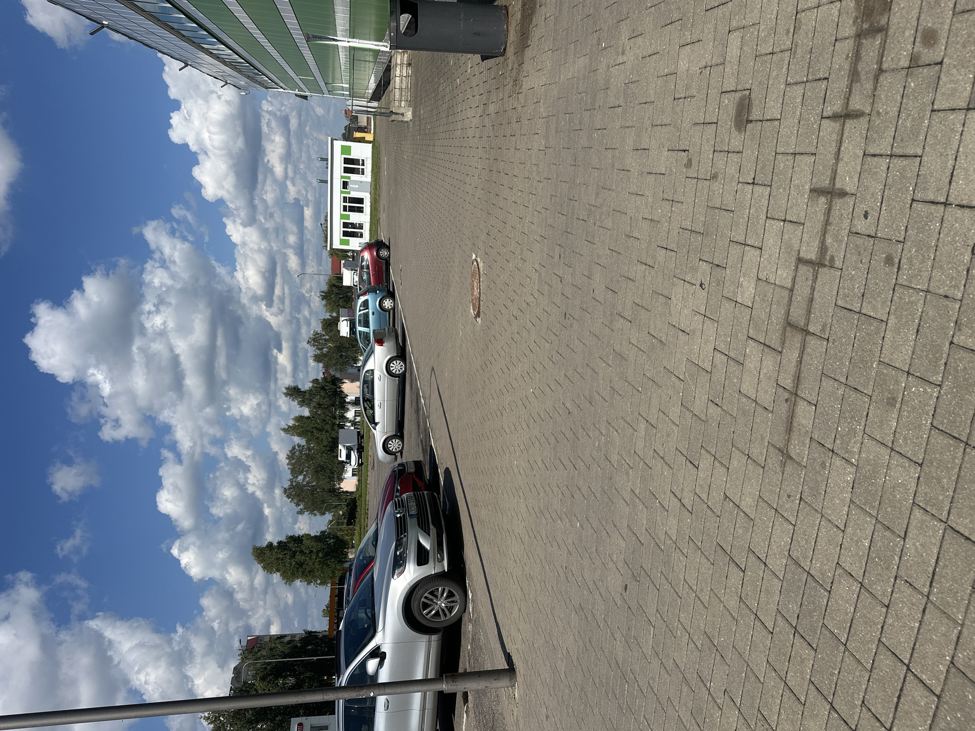 This image shows a parking lot paved with grey interlocking concrete pavers. In the foreground, the pavers extend into the distance, with some faint water stains visible. A dark grey trash receptacle stands on the right side of the image, adjacent to a sloped concrete ramp leading up to a modern building with a green and white facade. Several cars are parked in the lot. On the left, a silver SUV is parked, partially obscured by a dark grey pole. Behind it, a red car is parked. Further into the lot, a silver sedan is parked. In the background, there are trees, a small white building with green trim and multiple windows, and a white truck. The sky is blue with fluffy white clouds. The scene appears to be located in the city of Maladzyechna, Belarus.
