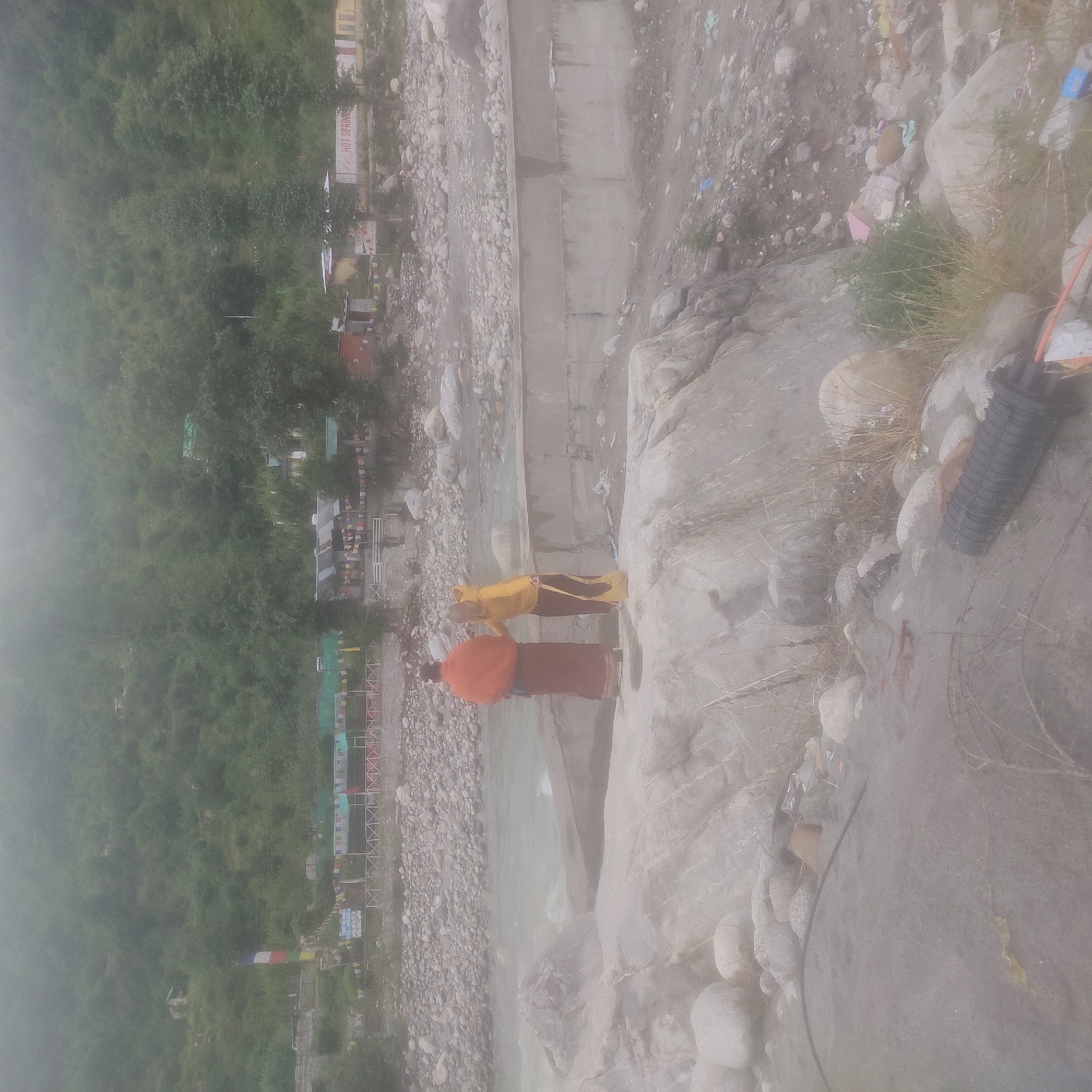 In the city of Kalath, India, two individuals dressed in monastic-style robes stand on a rocky shore beside a flowing river. The individual on the left wears an orange robe and a dark skirt, while the person on the right is in a yellow top and dark skirt. They are facing away from the camera, toward the river. The riverbank is strewn with numerous rocks and pebbles. In the background, a concrete wall lines the river, and beyond that, buildings and trees are visible on a misty, tree-covered mountainside. The area appears to be a roadside market or settlement, with various structures, some decorated with prayer flags, lining the landscape. The overall scene is hazy, suggesting misty or foggy weather conditions. The image captures a moment of pause or contemplation by the river in a mountainous, possibly rural, setting.