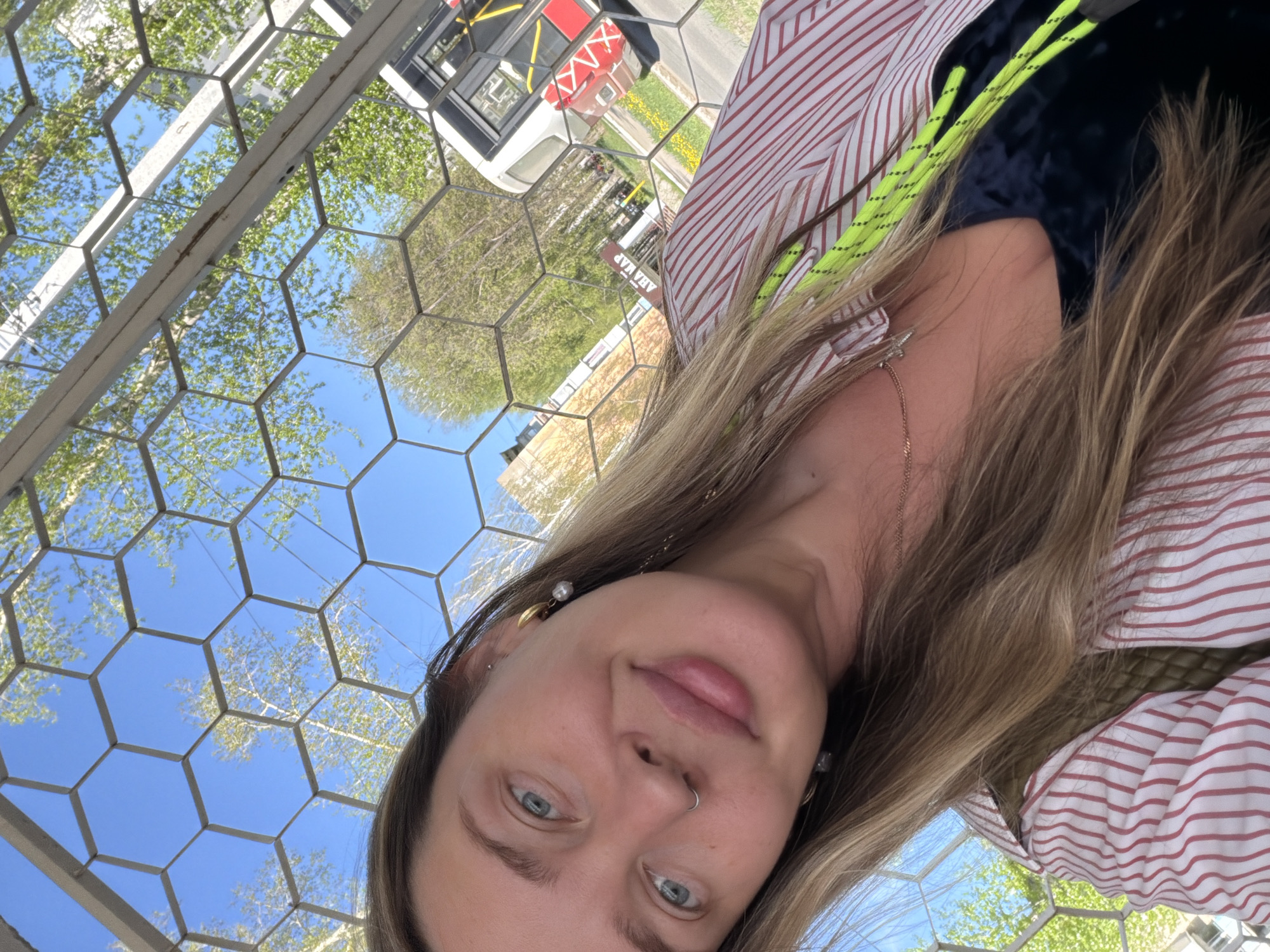 A selfie of a woman with blonde hair, wearing a striped shirt, taken outdoors under a metal structure with hexagonal openings that reveal a view of trees, sky, and a building in the background. The woman has light eyes, fair skin, and a nose ring. Behind the metal structure, part of a red and white bus is visible. The location is Chaikovsky, Russia.