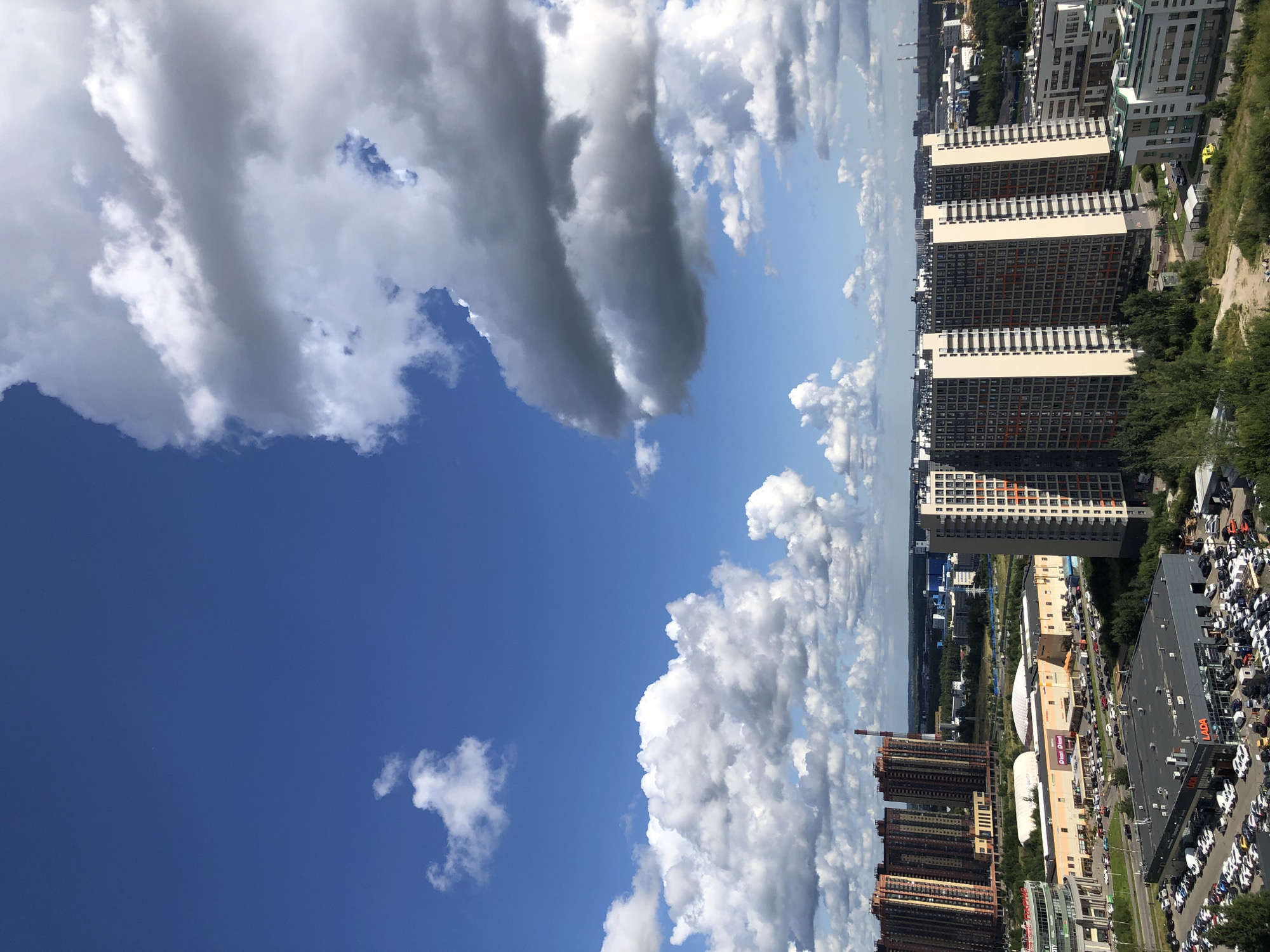 This is a high-angle, full shot of a city landscape on a sunny day. The sky is a vibrant blue with scattered white and gray cumulus clouds. In the foreground, there are several modern, high-rise buildings. To the left, a cluster of tall residential buildings with orange and brown facades stand prominently. In the center, a row of lighter-colored apartment buildings with numerous windows and some orange accents dominates the middle ground. To the right, another tall apartment building with a beige facade and orange vertical stripes is visible. Below these buildings, a commercial area is apparent, with a large building that appears to be a shopping mall, identified by signage with "Lenta" visible. A large parking lot is in front of the mall, filled with numerous cars. Further in the foreground, a car dealership is visible, with "LADA" branding and rows of cars parked outside. The surrounding area is dotted with trees and smaller buildings, with a distant view of more urban development and industrial structures on the horizon. The scene suggests a suburban or outer-city residential and commercial area. There are no people visible in the image.