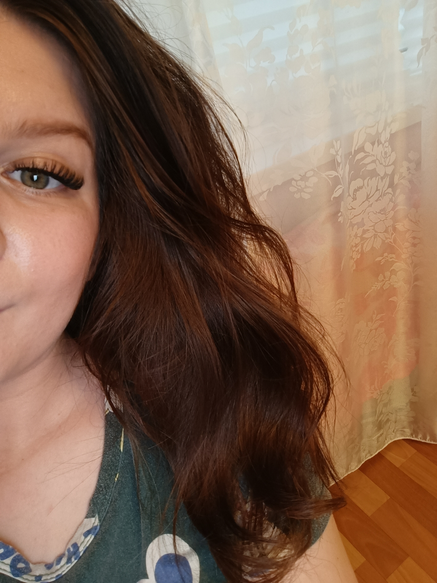 A person with auburn-brown hair is shown in a close-up shot. The person's left eye, adorned with dark, extended eyelashes, is visible. The person's hair is styled in loose waves, cascading toward the right side of the image.
The person is wearing a green T-shirt with a white and blue floral pattern around the neckline. A floral patterned, sheer curtain is visible in the background. The lower right corner of the image shows a wooden floor with a rectangular pattern.
There is no indication that the image was taken in Tobolsk, Russia.