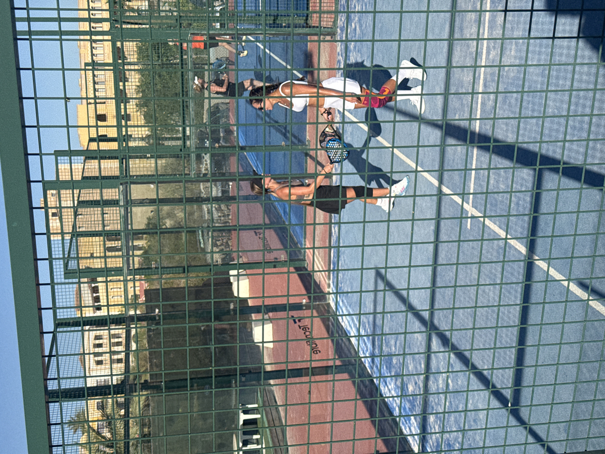 The image depicts an outdoor padel tennis court in Benidorm, Spain, viewed through a green chain-link fence. Two women are engaged in a game of padel tennis on the blue court. The woman on the left wears a dark top, a black skirt, and white athletic shoes, holding a padel racket. The woman on the right is dressed in a white athletic top and white shorts with pink knee pads, also holding a padel racket. In the background, other players can be seen on adjacent courts, and beyond them, buildings with yellow facades are visible, characteristic of the architecture in Benidorm. The sun casts shadows across the court, indicating a sunny day. The overall scene captures a moment of recreational activity in a sports facility.