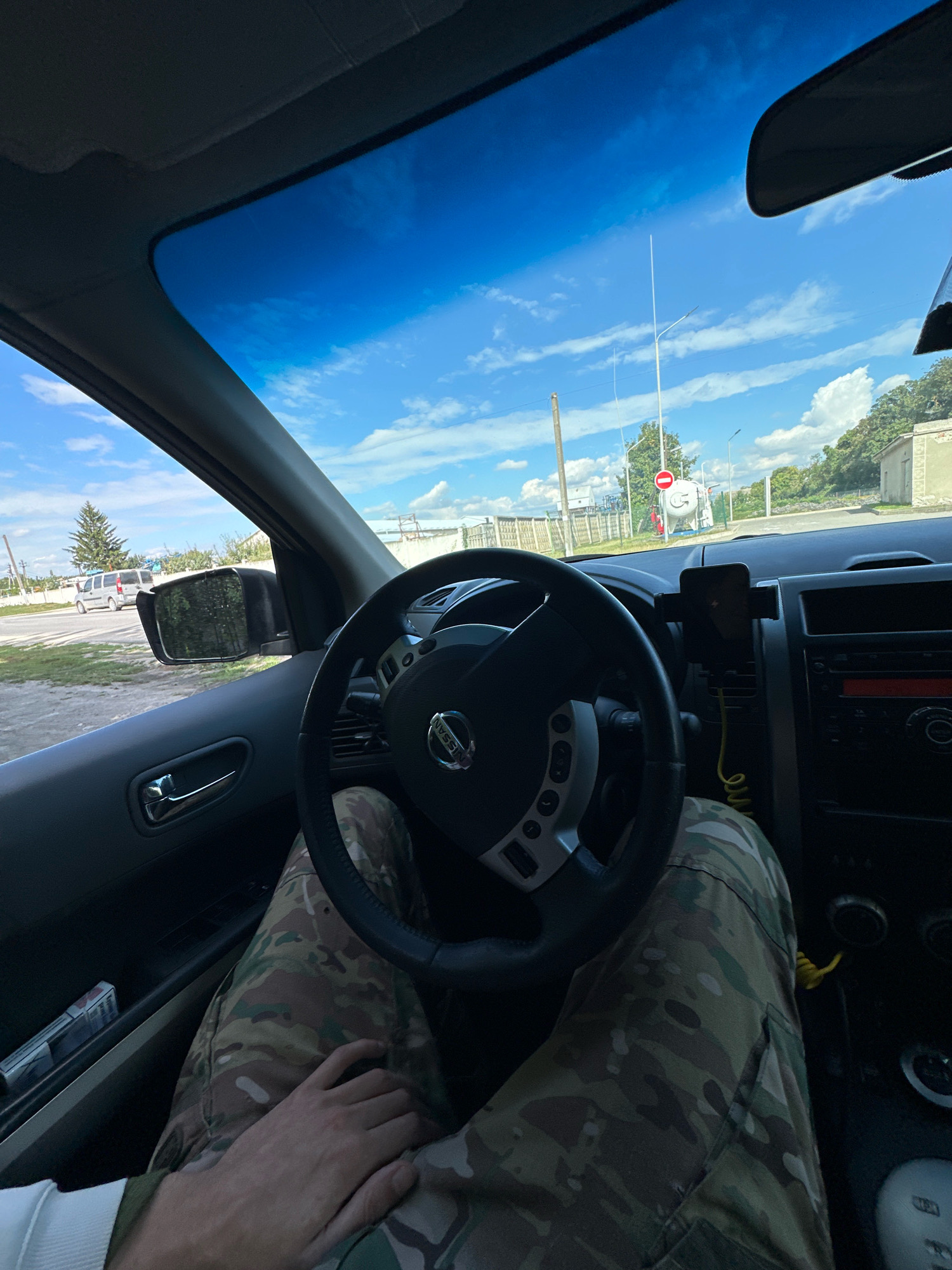 A POV shot from inside a vehicle captures the driver's hands and legs, clad in camouflage pants, resting on the steering wheel and dashboard. The car is parked on a dirt road in the city of Shatava, Ukraine, with a bright blue sky and scattered clouds visible through the windshield. The environment suggests a rural or semi-rural setting, with trees, a fence, and buildings visible in the background. A red and white traffic sign is visible in the distance.
