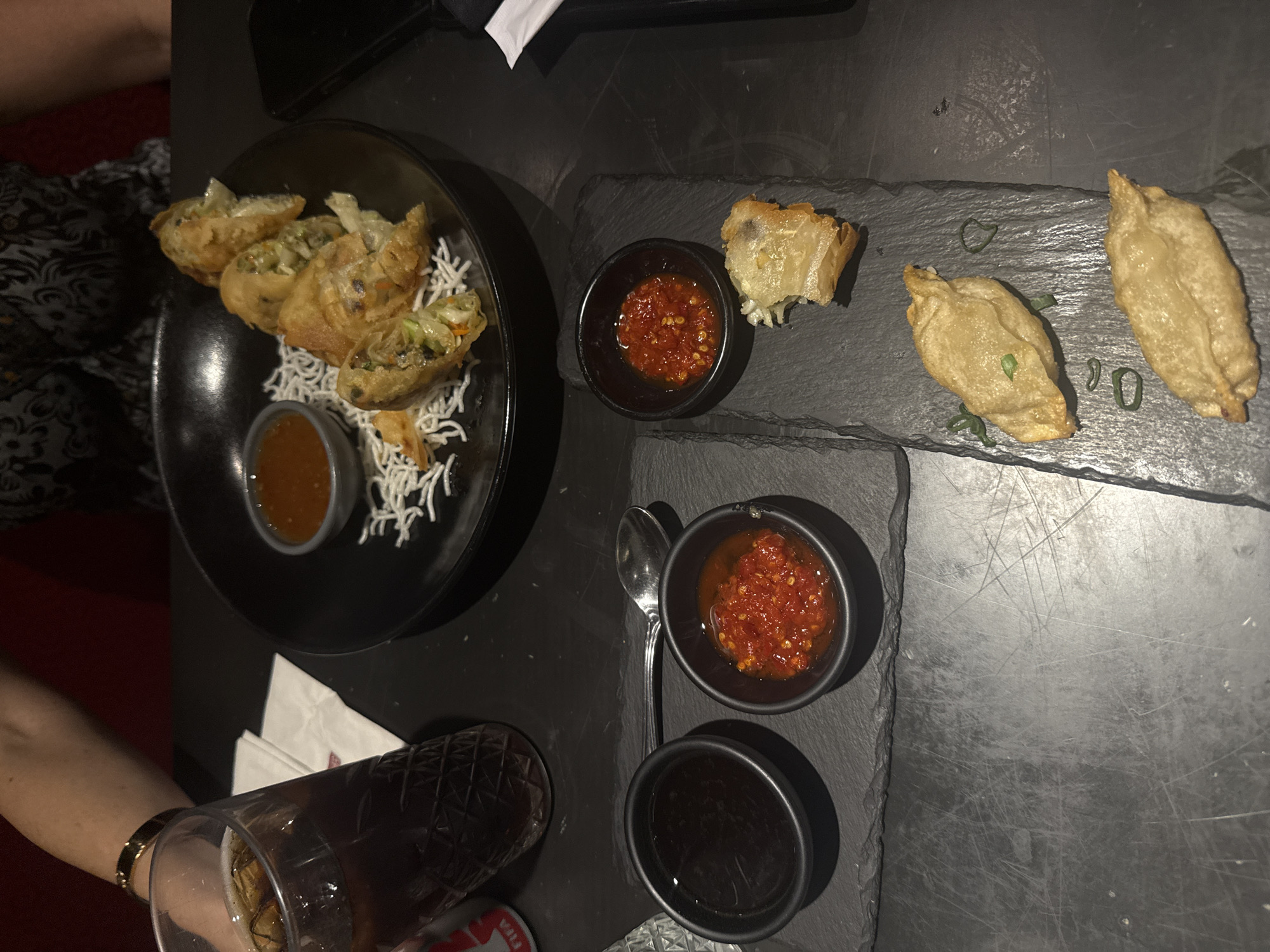 The image shows a table setting at a restaurant. On the left is a black bowl with fried spring rolls served on a bed of white crispy noodles. A small sauce bowl is also within the bowl. Next to it, two black slate plates hold Asian-style dumplings and small black bowls with dipping sauces. A glass with a dark beverage is visible in the lower-left corner. The scene appears to be indoors at a restaurant, possibly located in Mexico City, Mexico. The lighting is dim, creating a contemporary ambiance. A person's arm and the edge of a red seat can be seen in the bottom left corner.