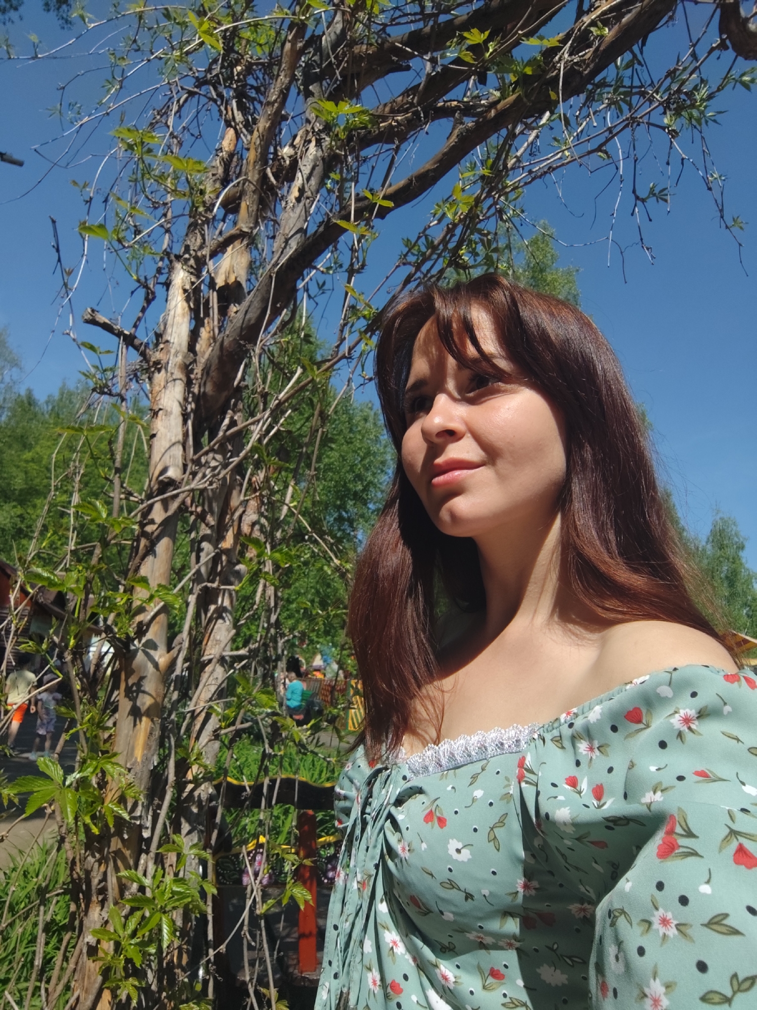 A woman stands outside on a sunny day in what appears to be a park setting. She has long brown hair with bangs, and she is wearing a light green off-the-shoulder dress with a floral pattern. The background includes a bare tree with a few new leaves budding. Further in the background there are other trees and hints of structures with a few people walking in the distance. The location is the city of Chaikovsky, Russia.