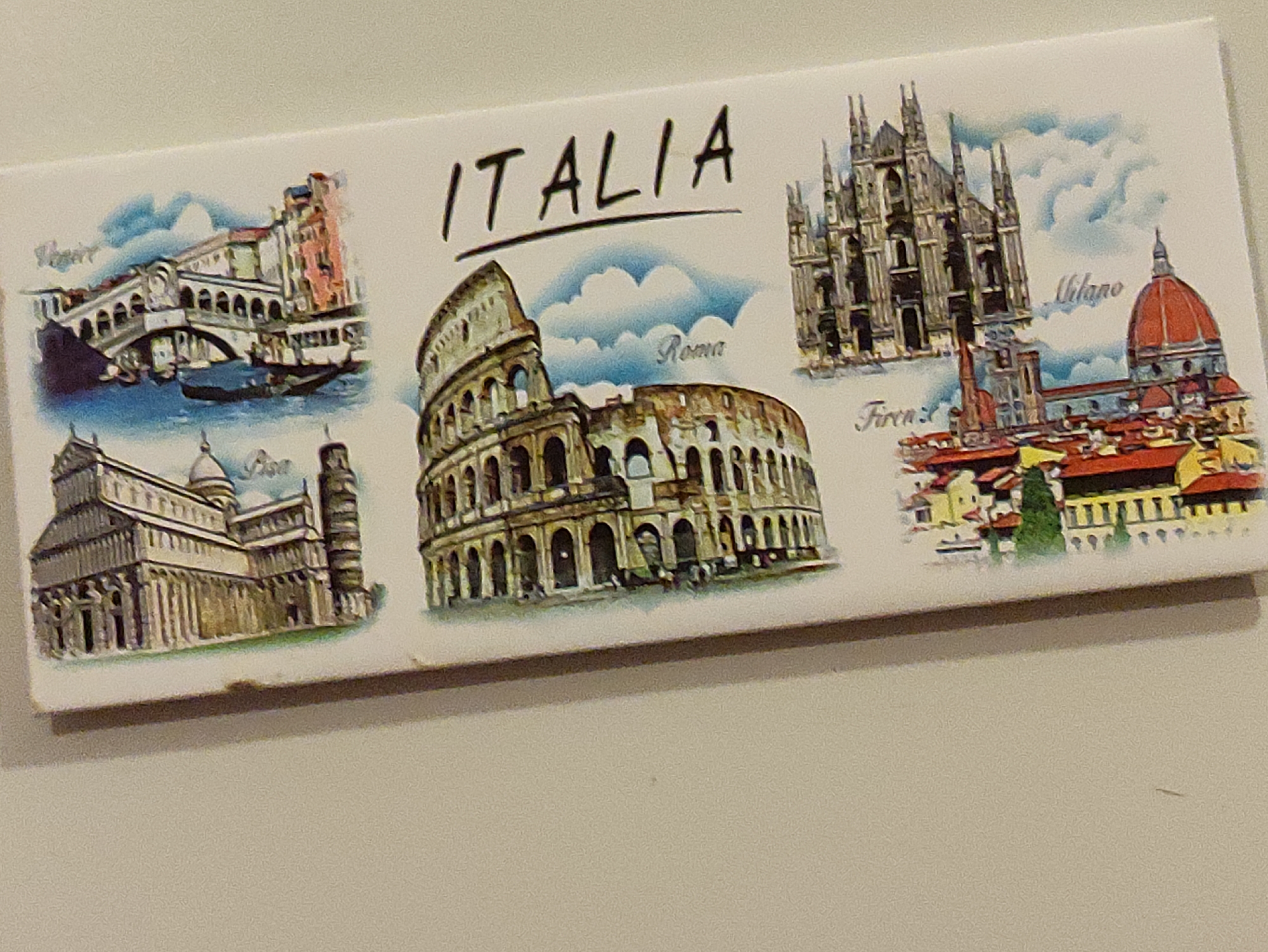 The image is a postcard or souvenir depicting famous Italian landmarks. In the top center, the word "ITALIA" is written. The postcard is divided into sections, each illustrating a different Italian city:
On the left, a bridge resembling the Rialto Bridge in Venice is shown with a gondola on the water. The text "Venice" is visible. Below this, the Leaning Tower of Pisa and a cathedral are depicted, with the text "Pisa".
In the center, the Colosseum in Rome is prominently featured, with the text "Roma" written above it.
On the right, the Duomo di Milano (Milan Cathedral) is illustrated, with the text "Milano" above it. Below this, a scene of Florence is shown, featuring buildings with red roofs and a dome, and the text "Firenze".
The background of the postcard features a light blue sky with white clouds. The overall style is illustrative and colorful. The postcard is positioned on a light-colored surface, possibly a table. The location of Krushev Dol, Bulgaria is not depicted or relevant to the content of the image.