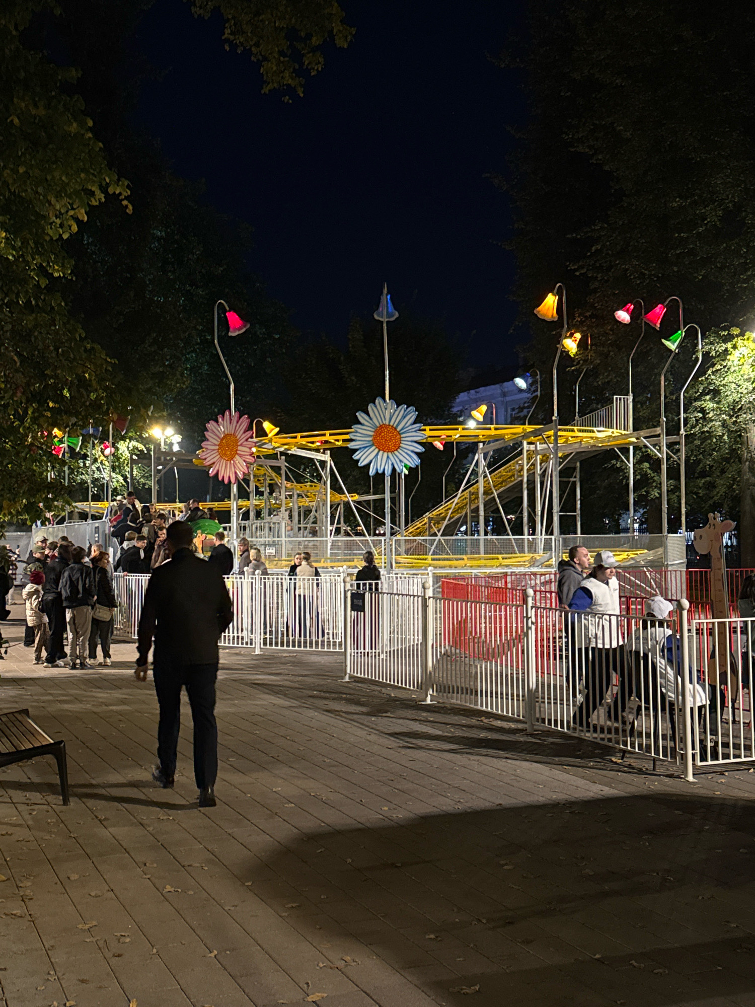 The image depicts an outdoor amusement park scene at night. A yellow roller coaster with a gentle slope is visible in the background, decorated with large daisy-like floral elements. Several people are gathered in the area, some appearing to be in line for the ride, while others are standing and talking. The park is illuminated by various decorative lights, including red, yellow, and green bell-shaped lamps mounted on poles, as well as string lights. A wooden walkway with scattered autumn leaves is in the foreground, and a dark park bench is positioned to the left. A white metal fence encloses a section of the park. The overall atmosphere suggests a festive evening at an amusement park in the city of Tver, Russia.