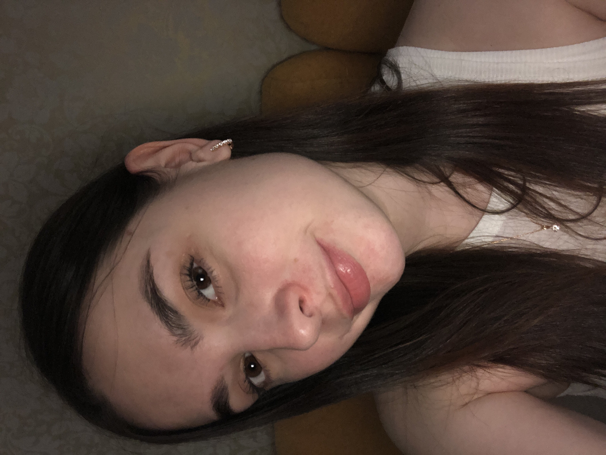 A close-up, slightly angled shot features a woman's face. Her complexion is fair, and she has dark brown hair. Her eyes are open, and her lips are slightly parted. She is wearing a white top. Her earring is visible in her left ear. The background is blurry, suggesting a soft, diffuse lighting environment. There is no verifiable information about the location being in Noviy, Russia, or any specific city.