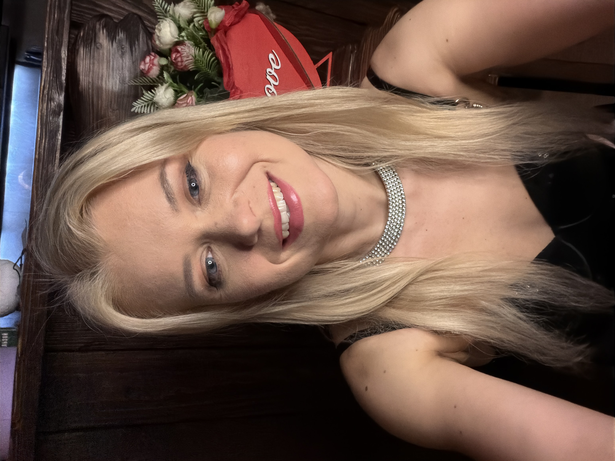 A close-up, selfie-style photograph shows a fair-skinned woman with blonde hair and blue eyes smiling at the camera. She is wearing a black sleeveless top and a sparkly silver choker necklace. Her makeup includes pink lipstick and eyeshadow. In the background, a wooden wall and shelf are visible. On the shelf, there is a bouquet of pink and white flowers arranged in a red heart-shaped box that says "Love." The lighting is dim, suggesting an indoor setting, possibly a restaurant or a home, and the background elements do not provide specific clues to its location within Sochi, Russia.