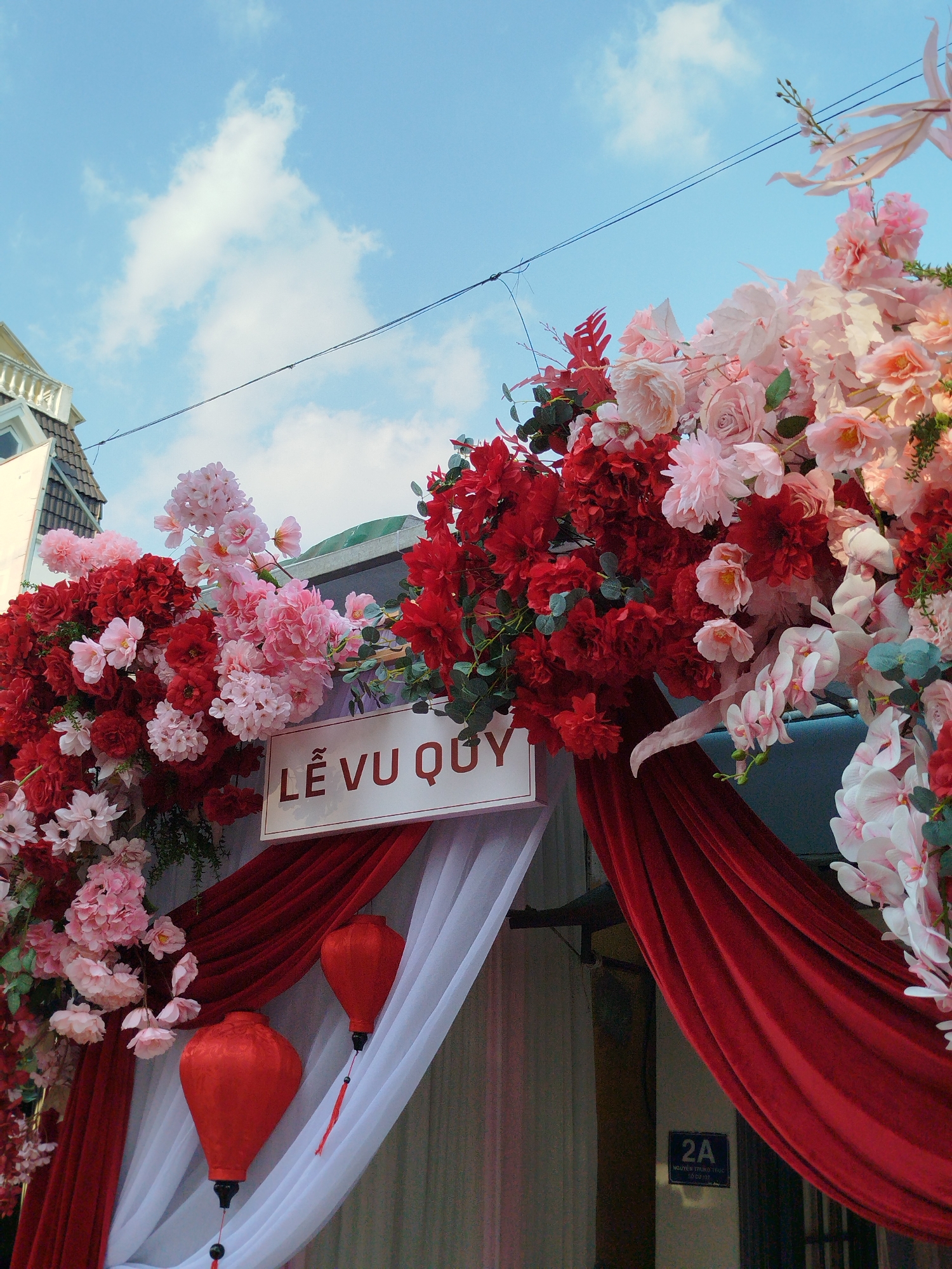An outdoor scene captures a decorated entrance under a partly cloudy blue sky. The entrance features an archway or frame heavily adorned with artificial flowers, primarily in vibrant red and various shades of pink, including roses, hydrangeas, and orchids, augmented by green foliage. Red and white fabric drapes are arranged at the entrance, with two red, teardrop-shaped lanterns suspended against the white fabric. A central white rectangular sign with a brown border displays "LỄ VU QUY" in black capital letters. In the upper left, a portion of a building with a brown tiled roof and a white balustrade is visible. Two black utility wires span the upper middle of the frame. On the lower right, a blue street sign is affixed, clearly indicating "2A" and "NGUYỄN TRUNG TRỰC, P.1 - ĐÀ LẠT," pinpointing the location as 2A Nguyễn Trung Trực Street, Ward 1, Da Lat, Vietnam. No individuals are present in the image.