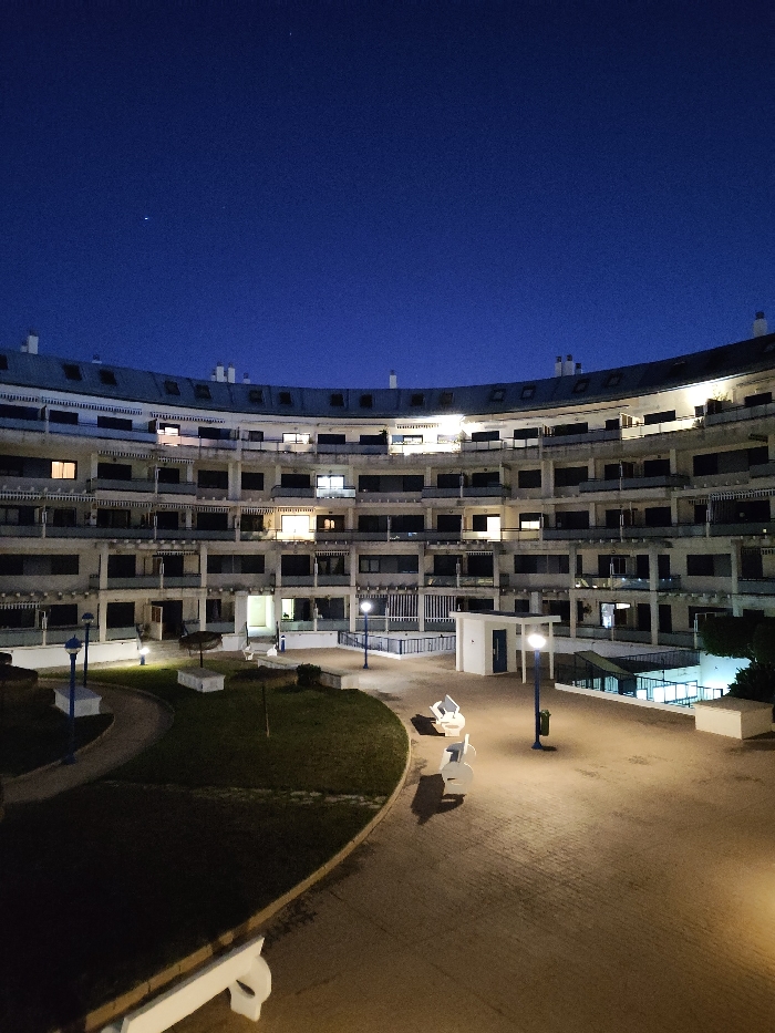 A multi-story, curved apartment building faces an open courtyard. Balconies with dark windows line each level. Lights shine from several apartments. A paved area fills the courtyard, illuminated by tall street lamps. Grass borders the pavement. Benches are scattered throughout the area. The sky is a deep blue, indicating it is nighttime.