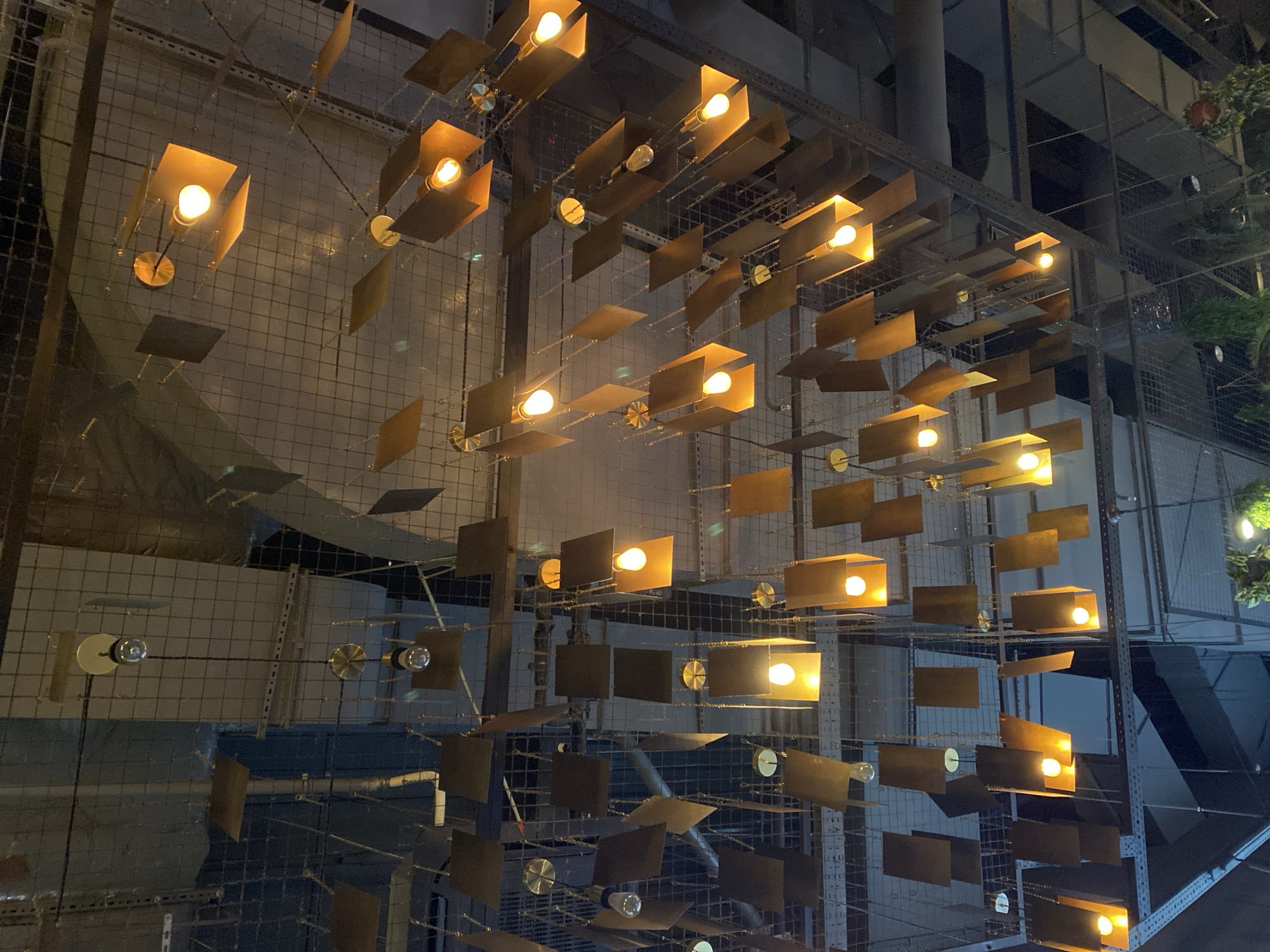 The image shows a complex lighting fixture consisting of multiple rectangular shades and exposed light bulbs suspended from a wire mesh ceiling. The shades are metallic, possibly bronze or copper colored, and appear to be arranged in a somewhat random, yet structured, pattern. The location appears to be an interior space with industrial or modern architectural features, potentially a restaurant, retail space, or art installation. The wire mesh ceiling is suspended below a high ceiling, possibly in a warehouse-style building. In the background, there is an element suggesting a staircase, and a glimpse of foliage, possibly indoor plants. The overall impression is one of a deliberately designed space with a focus on lighting as a decorative element. The image cannot be pinpointed to Yekaterinburg, Russia, with the visual information provided.