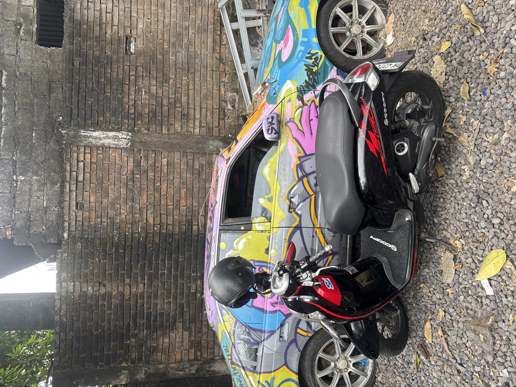 The image shows a black scooter parked in front of a car that is covered in colorful graffiti. The car is a sedan with silver rims and a broken side mirror. The scooter is a Honda Scoopy, also with silver rims, and has a black helmet resting on its handlebars. The scene appears to be an outdoor area with a gravel ground and a brick wall in the background. The location is identified as Kuta Selatan, Indonesia. The environment looks somewhat unkempt, with fallen leaves and some debris on the ground. There are no people visible in the image.
